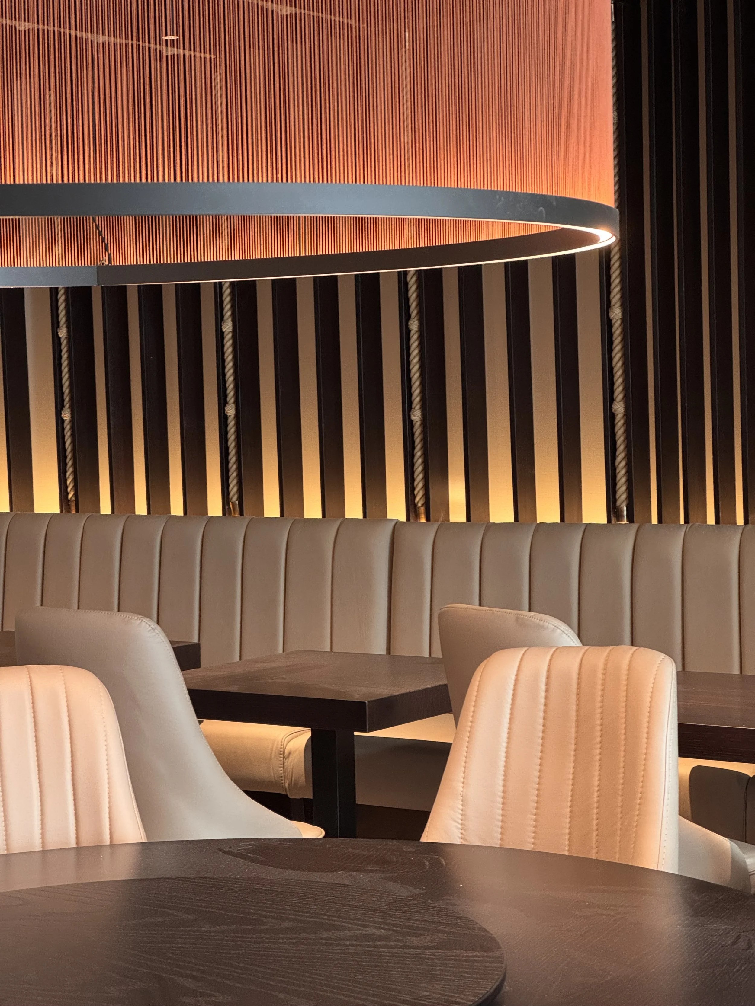 Modern restaurant interior with beige upholstered booths, dark wooden tables, and peach-colored chairs. A large red pendant light fixture hangs from the ceiling, and vertical black slats with rope accents decorate the wall.