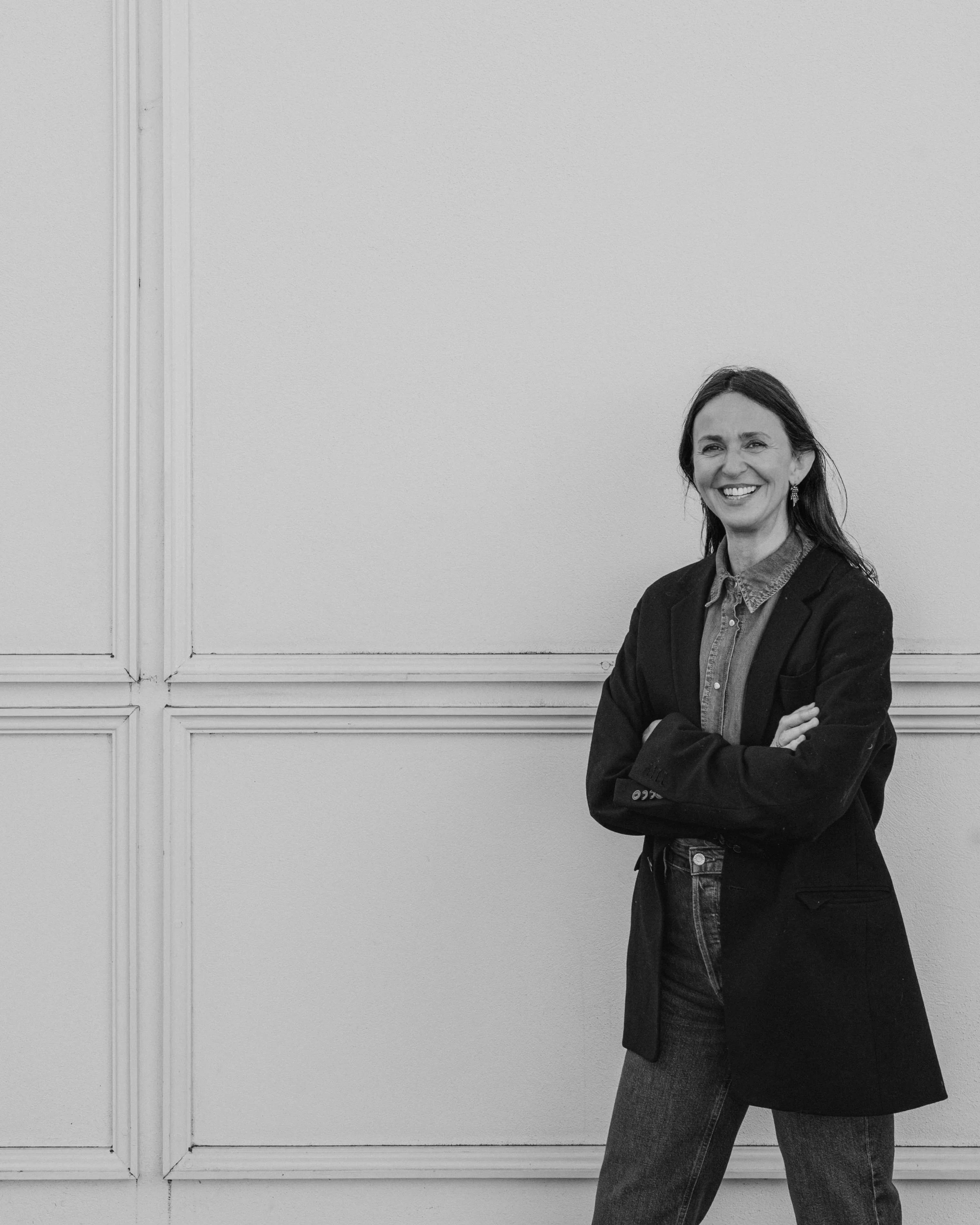 Smiling woman in denim shirt and black blazer standing against a cream-colored wall with arms crossed.