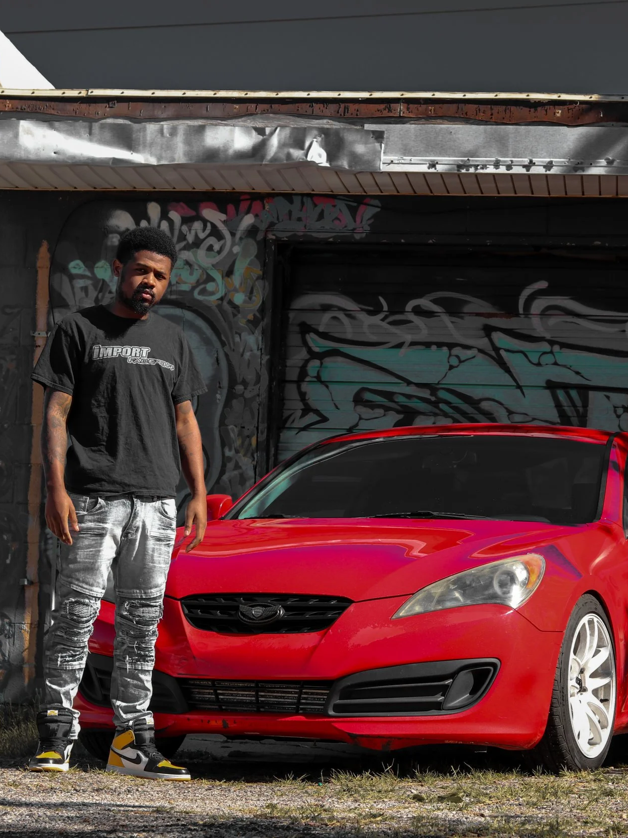 A man with a black T-shirt and distressed jeans standing next to a red sports car in front of a graffiti-covered wall.