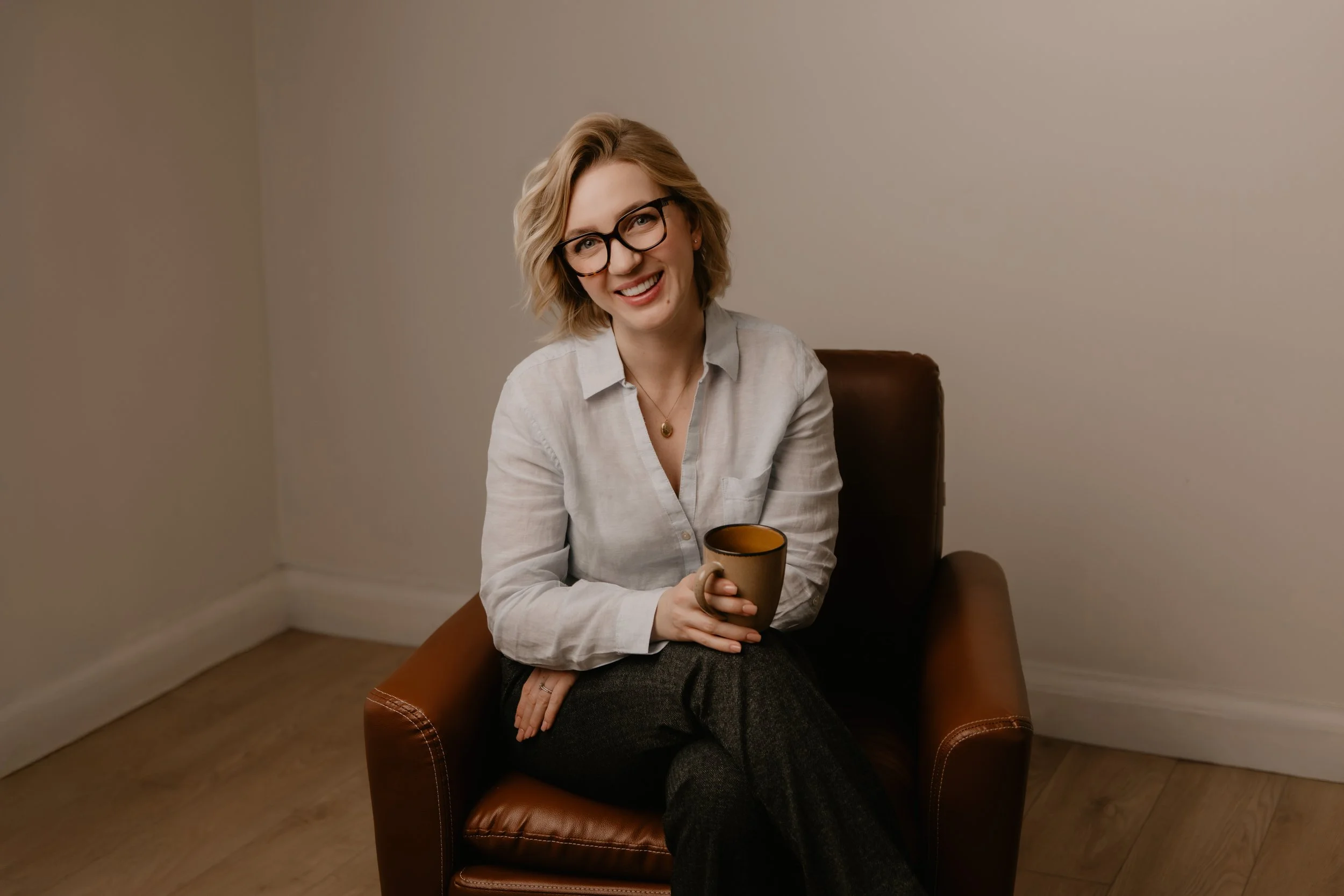 A woman with blonde curly hair and glasses sitting in a brown leather chair, holding a mug, smiling, in a plain room with beige walls and wooden flooring.