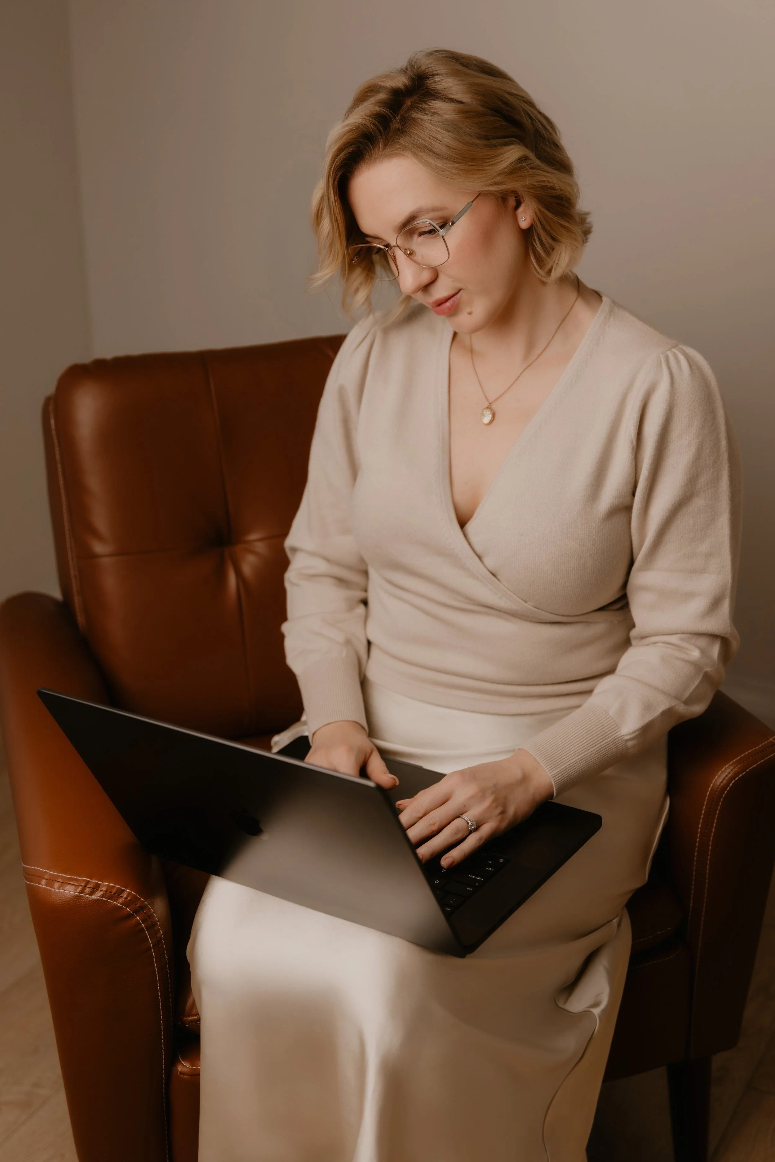 A woman with blonde hair and glasses sitting on a brown leather armchair using a black laptop.