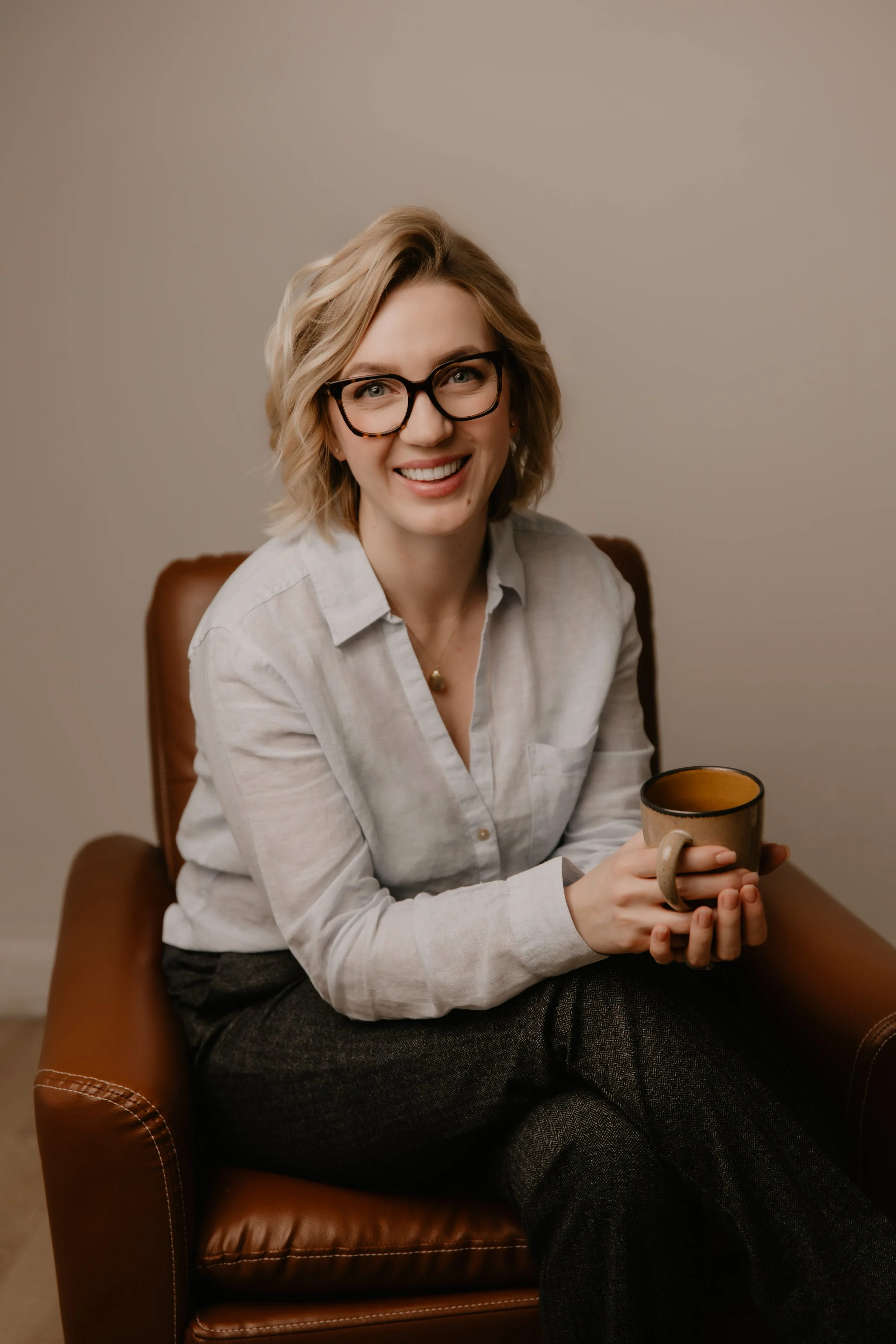 A woman smiling while sitting in a leather chair, holding a coffee mug, wearing glasses, a light gray shirt, and dark pants.