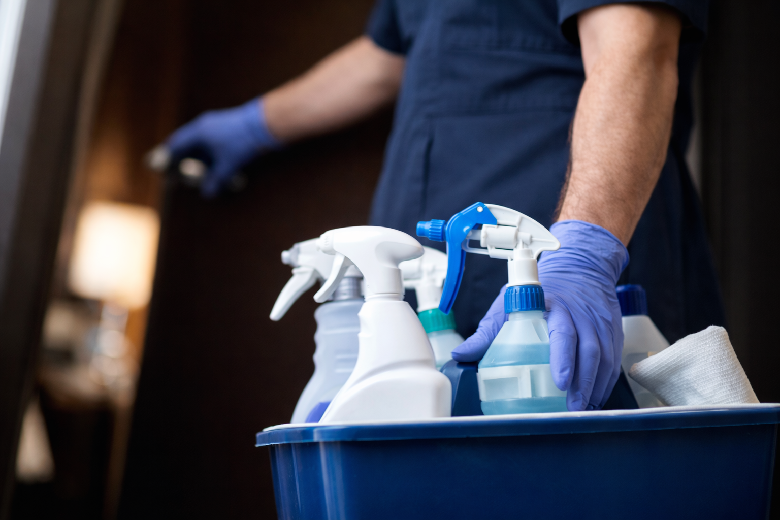 Person trägt blaue Handschuhe und hat eine Kiste mit Reinigungs- sprühflaschen und Tuch.