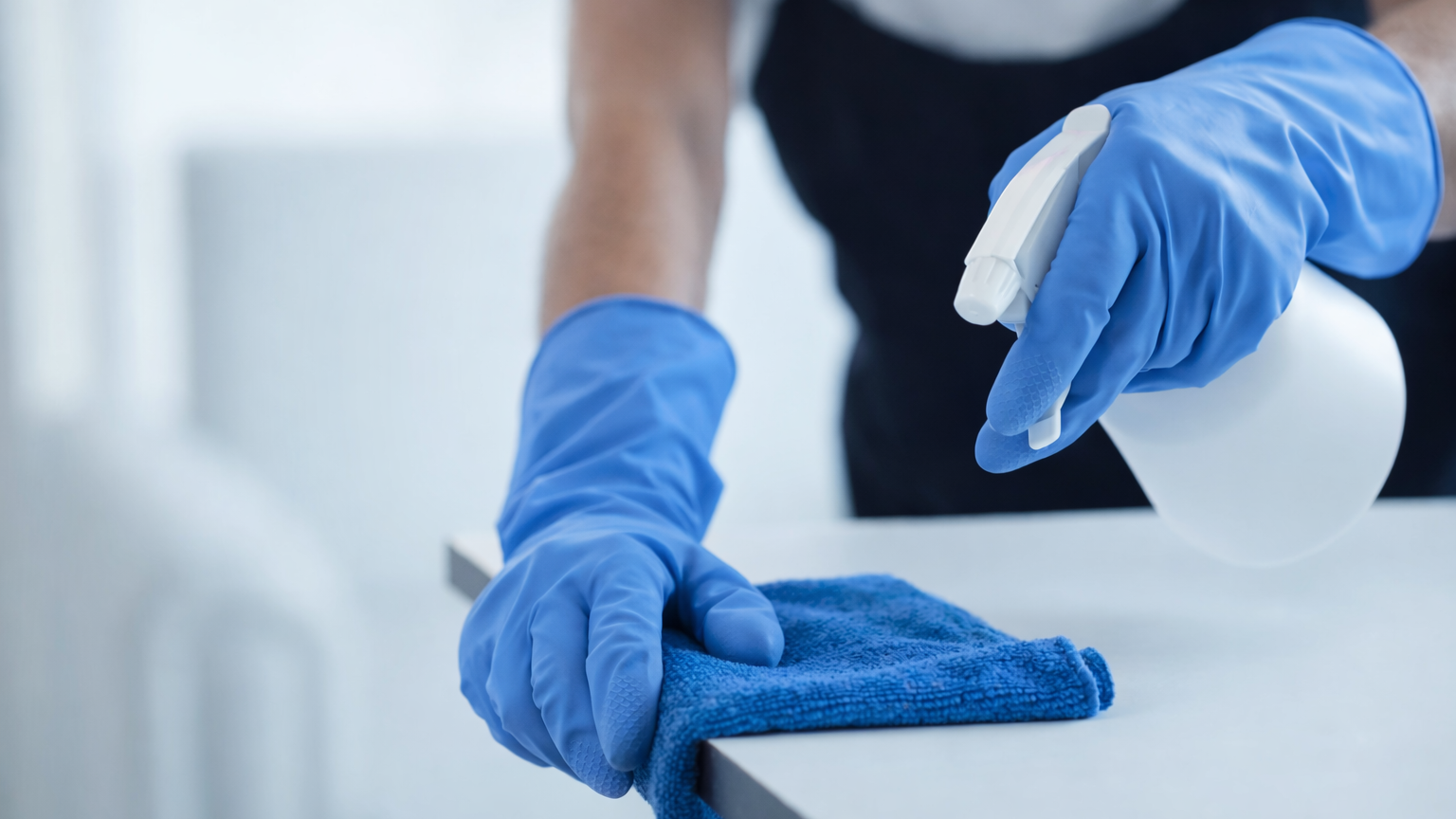Nahaufnahme von einer Person, die mit blauen Gummihandschuhen einen weißen Sprühreiniger auf einer weißen Oberfläche mit einem blauen Tuch aufträgt, vermutlich zur Reinigung.