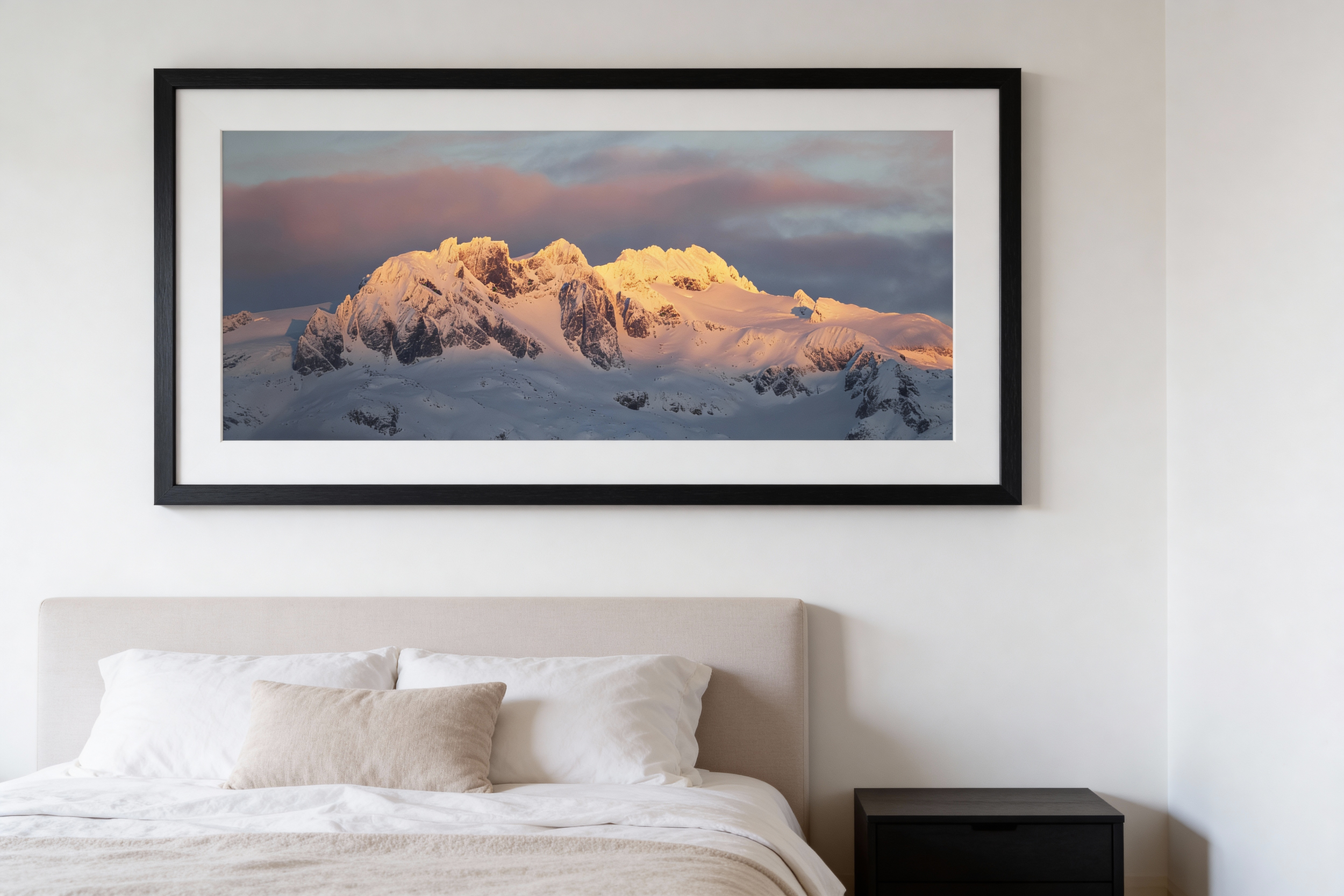 Framed landscape photograph of snow-covered mountain peaks during sunset hanging above a bed in a bedroom.