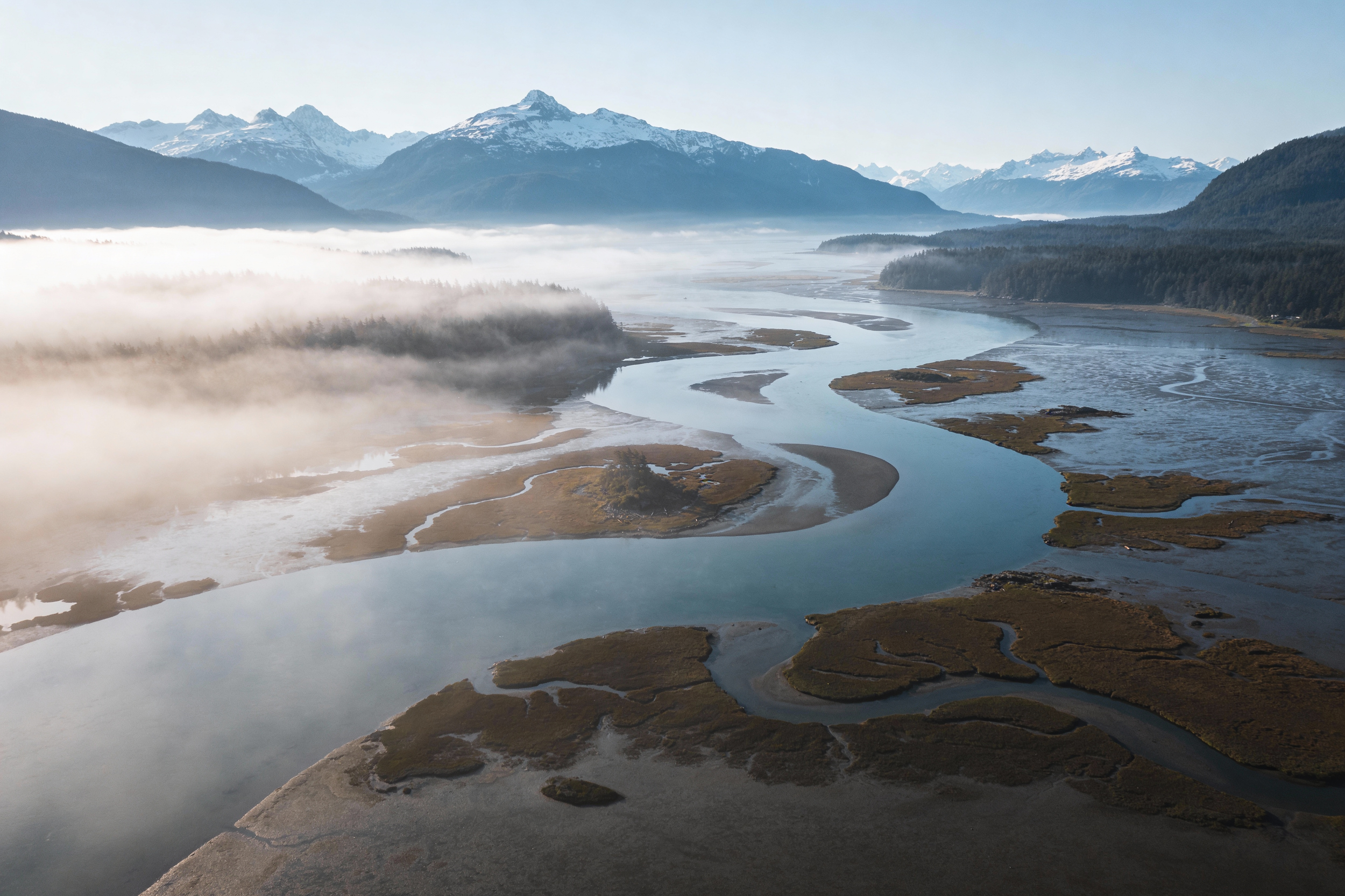 Aerial view of a river meandering through marshland with snow-capped mountains and morning fog near Revelstoke, British Columbia