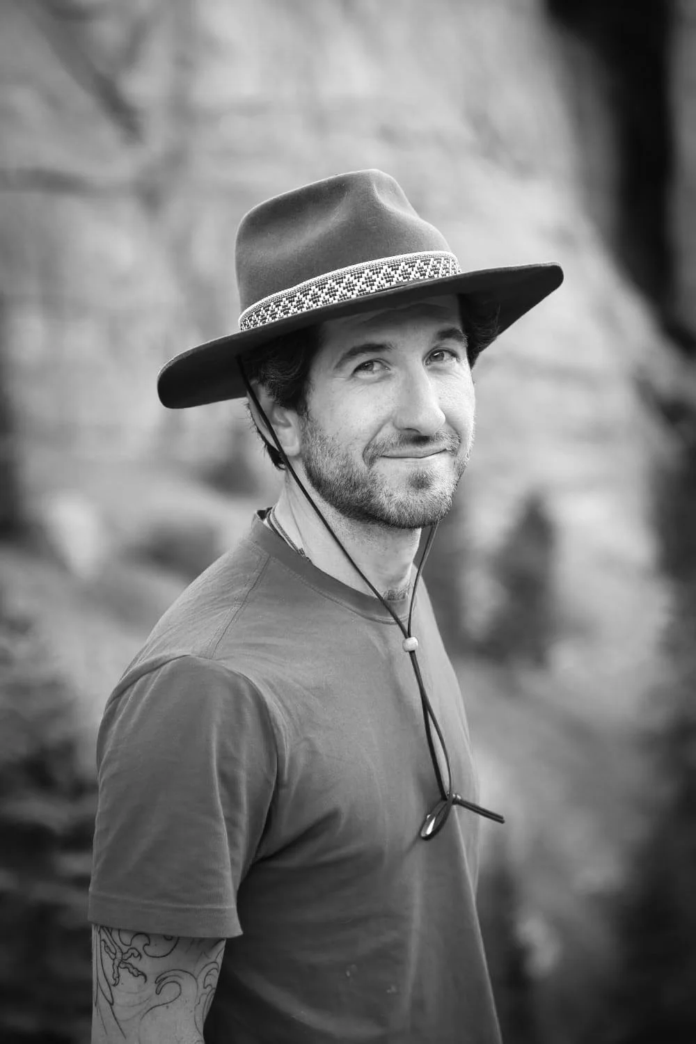 Black and white photo of a Kyle Smith wearing a wide-brimmed hat with a decorative band, t-shirt, and a tattoo on his arm, standing outdoors with a blurred natural background.