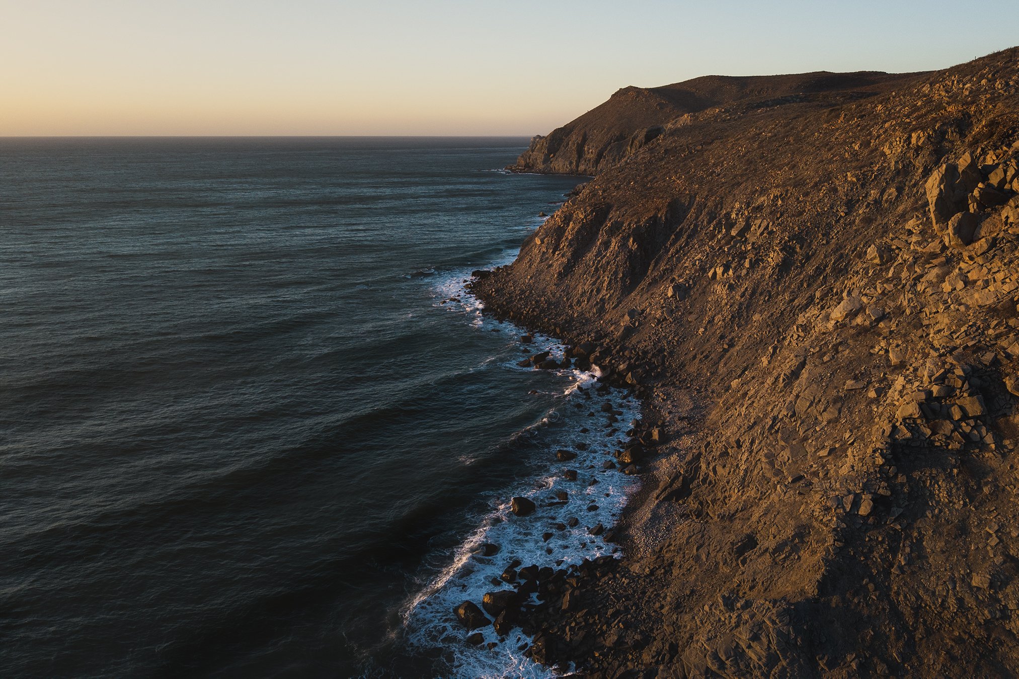 Aerial view of a rocky coastline during sunset with waves crashing against the shore.