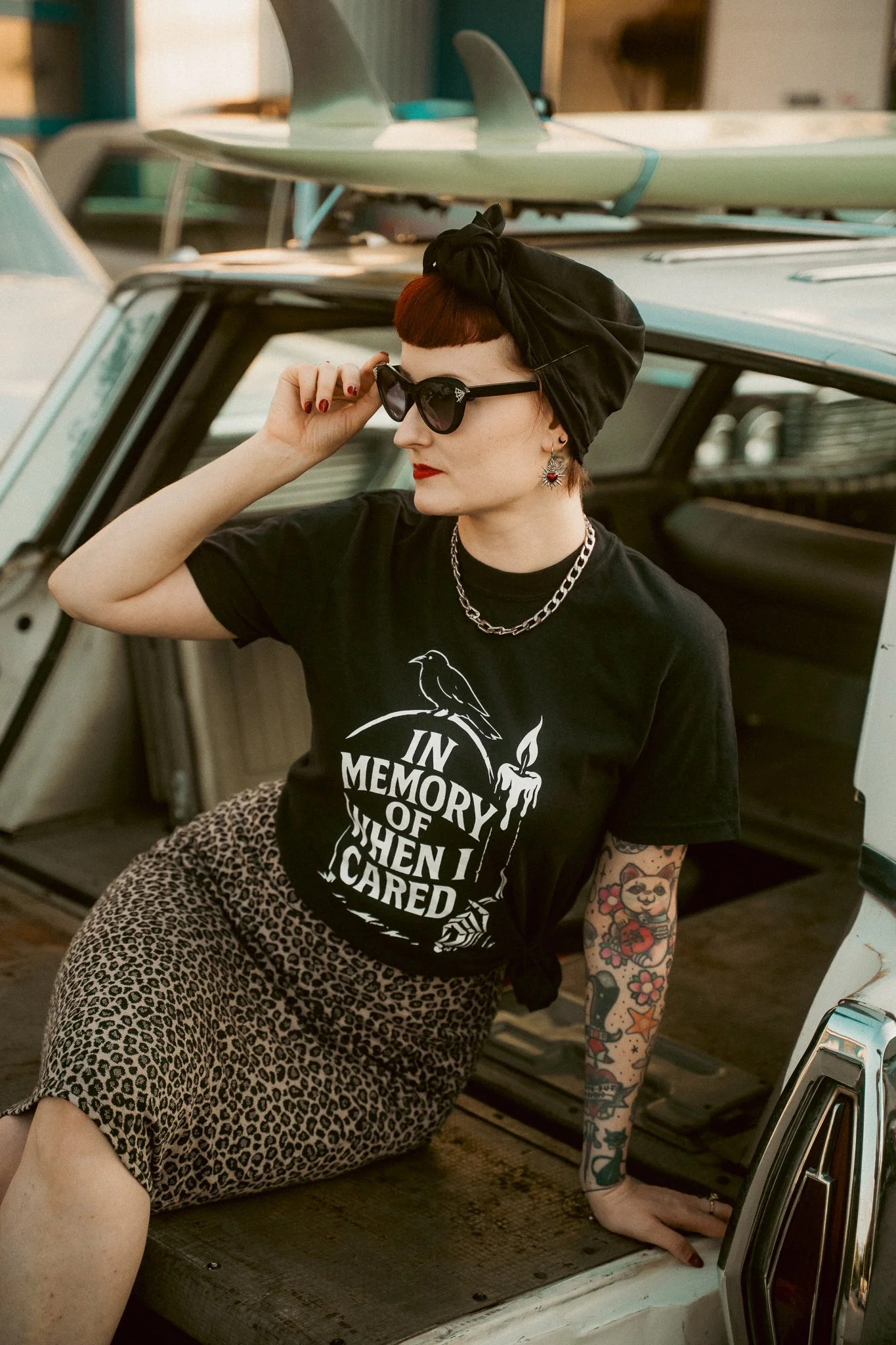 A woman with short hair, wearing sunglasses, a headscarf, a black graphic t-shirt, and leopard print pants, sitting on the floor with one hand on the ground, next to a vintage car with surfboards on top.