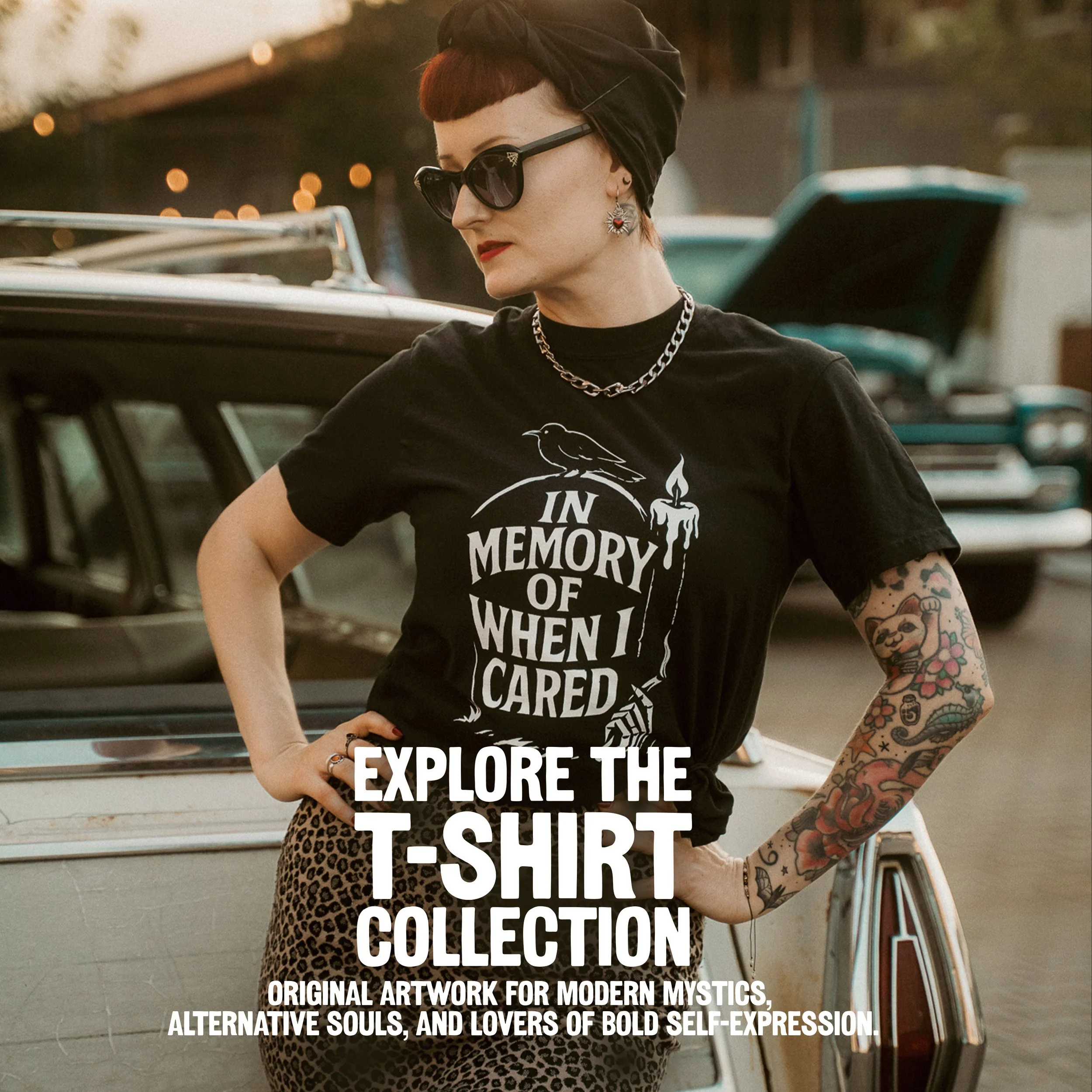 A person with short red hair wearing large black sunglasses, earrings, and a black headscarf, standing outdoors next to vintage bikes, wearing a black T-shirt with white text, with warm lighting and a blurred background.