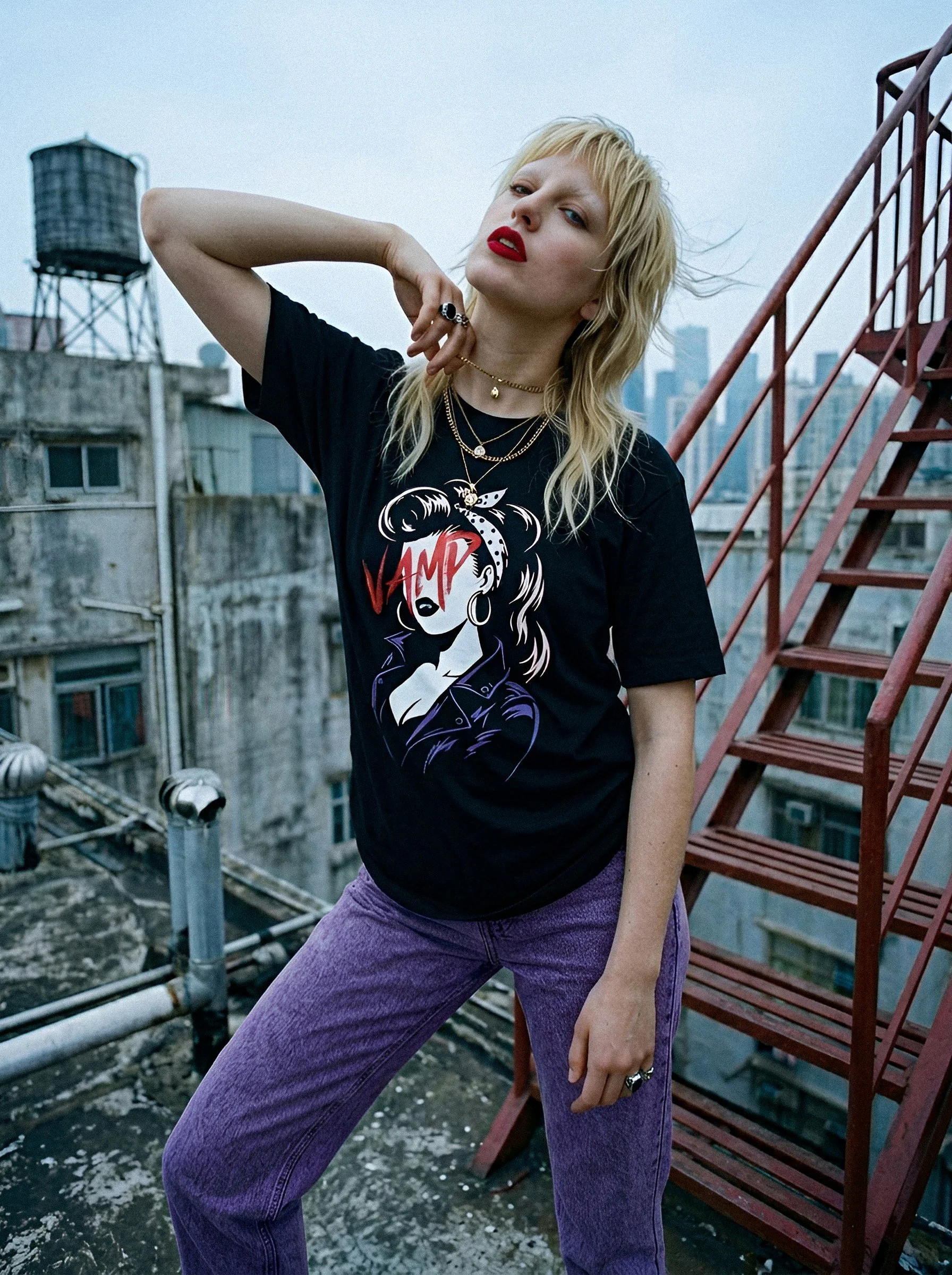 A woman with blonde hair, red lipstick, wearing a black graphic t-shirt and purple pants, posing on a rooftop with a city skyline in the background.