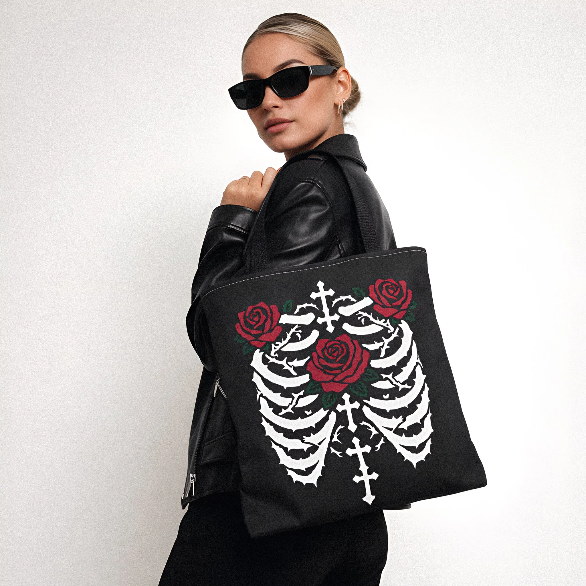 A woman wearing sunglasses and a black leather jacket holding a black tote bag with a white skull and bones, red roses, and crosses design on it.