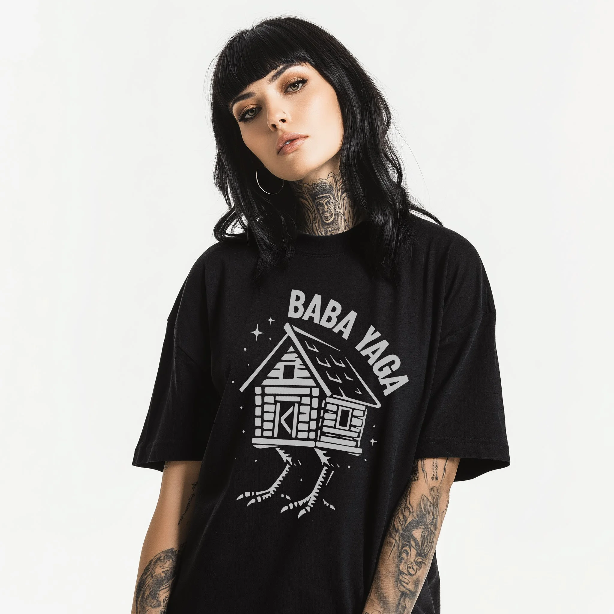 A young woman with black hair, tattoos, and hoop earrings wearing a black t-shirt with a graphic of a house and the words "BABA YAGA" on it against a plain white background.