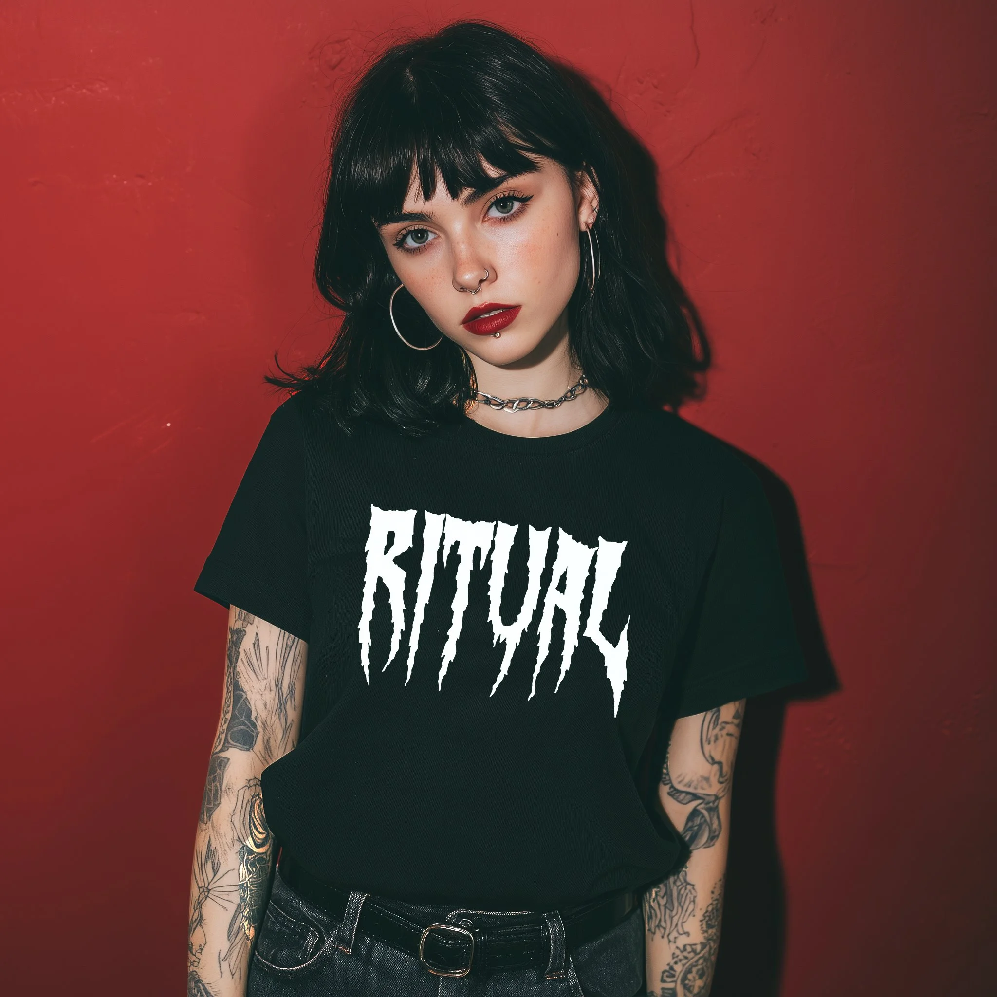 Young woman with dark hair and tattoos wearing a black 'BATWOLF' T-shirt, standing against a red wall.