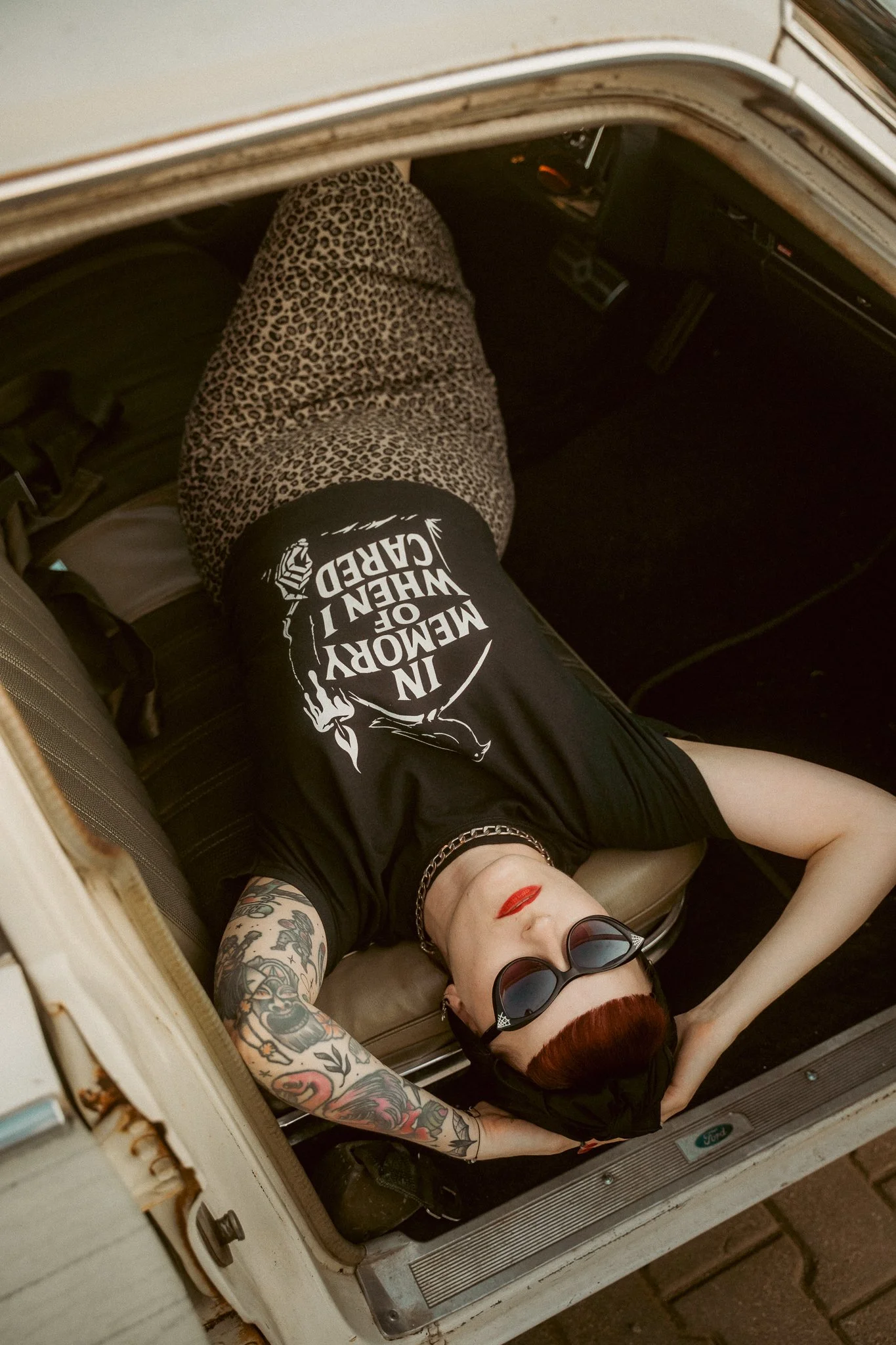 A person with red hair, tattoos on their arm, wearing sunglasses and a black T-shirt with white text, lying on the floor of an open vintage camper. The T-shirt has the phrase 'In Memory of W.W. HART CURED' on it, and they are wearing leopard print pants.