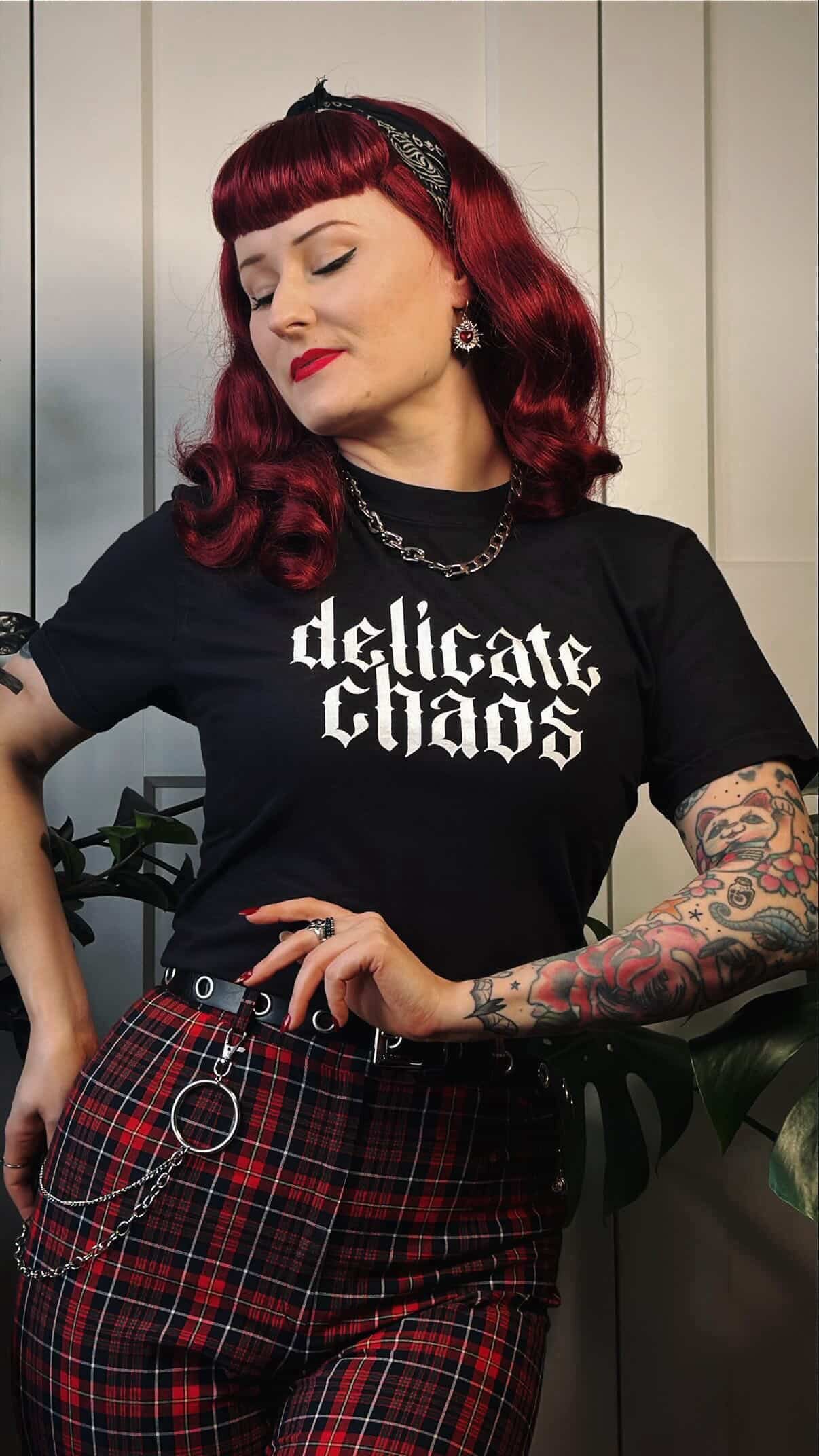 A woman with bright red hair styled in retro waves and a short blunt fringe, wearing a black headband, red lipstick, and winged eyeliner. She has tattoos on her left arm, including a colorful floral design and a cat face. She is dressed in a black T-shirt with the phrase 'delicate chaos' in gothic font, layered with silver jewelry, including a chain necklace, earrings, and rings. Her plaid skirt features red, black, and white patterns, accessorized with silver chains and rings.