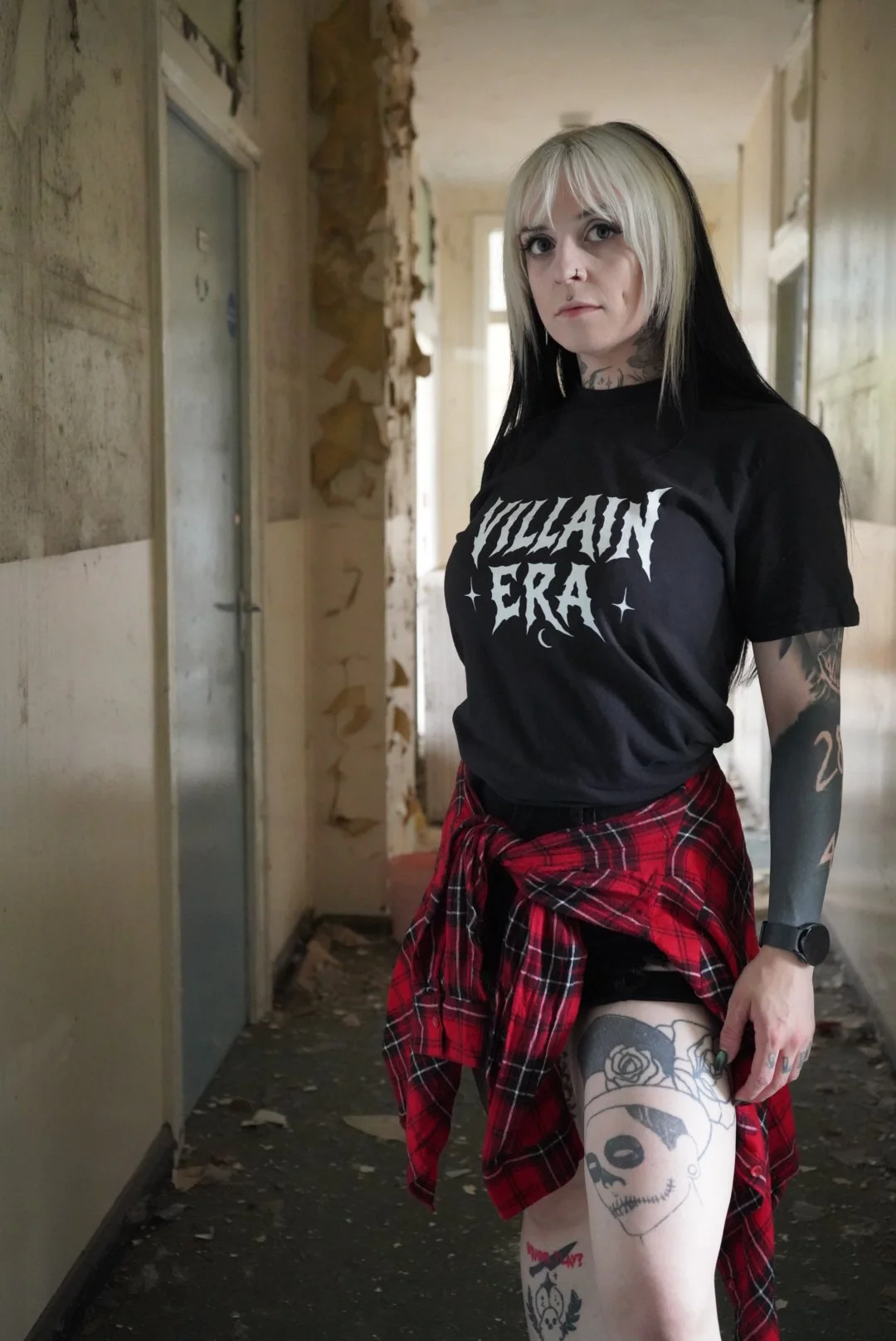 A woman with platinum blonde and black hair standing in a dilapidated, abandoned building, wearing a black T-shirt with 'VILLAIN ERA' printed on it, red plaid shirt tied around her waist, shorts, and showing various tattoos on her arms and legs.