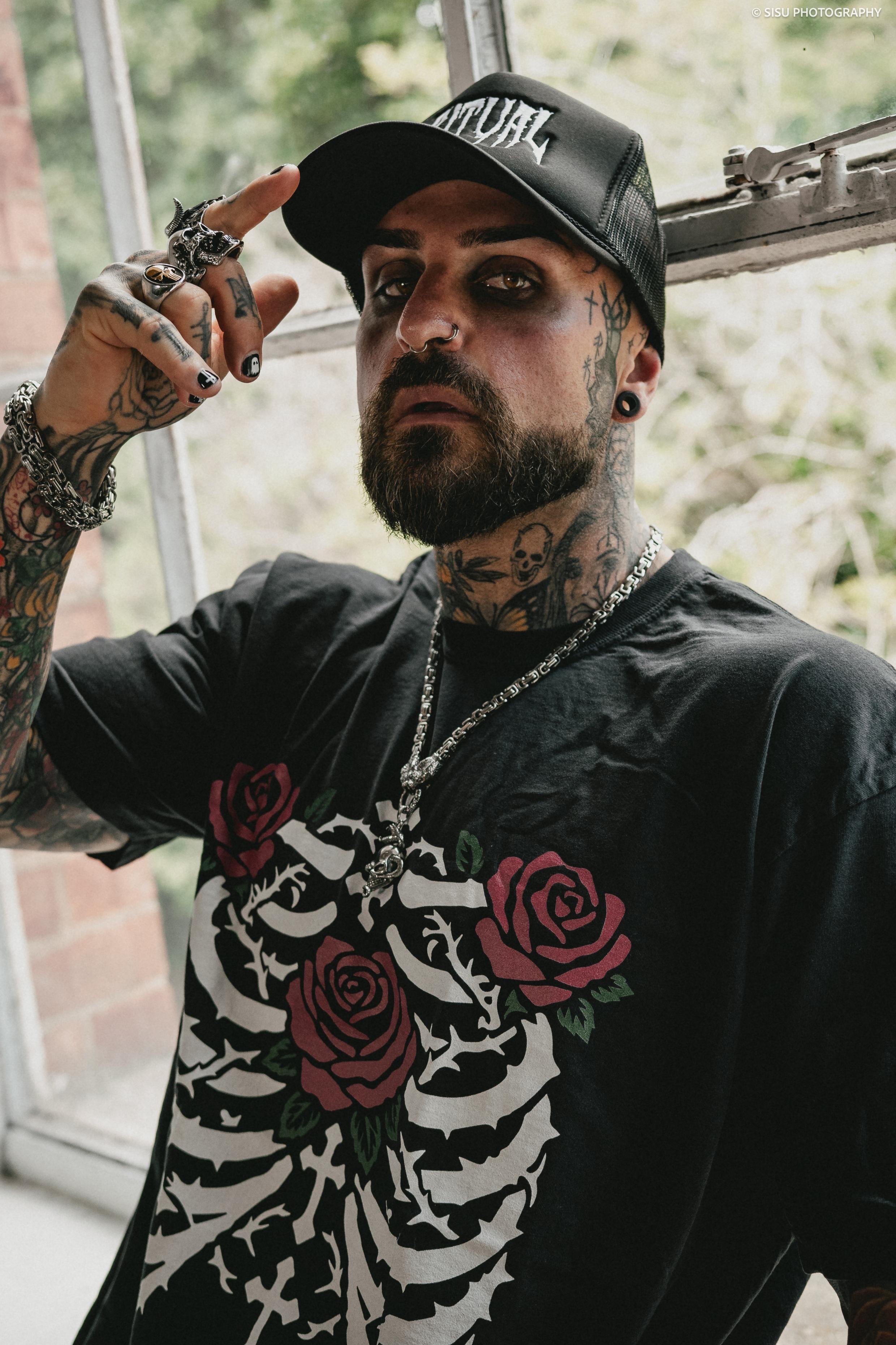 A man with tattoos on his face, neck, and arms posing indoors near a window. He is wearing a black cap, a black graphic T-shirt with roses, and Jewelry.