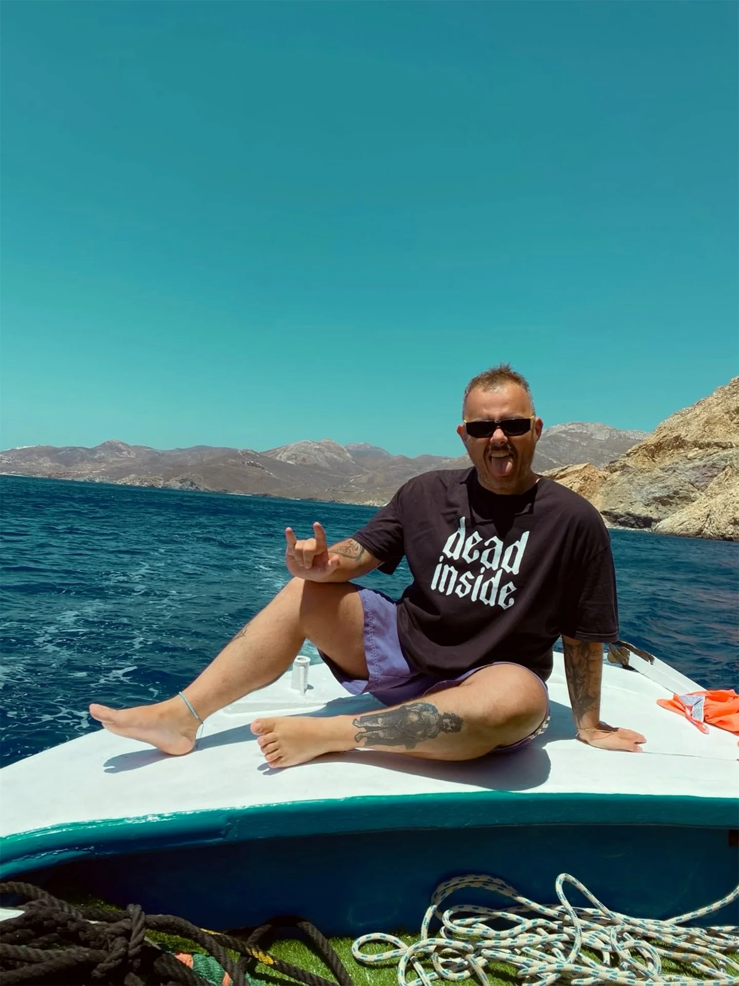 Man sitting on the edge of a boat, smiling with tongue out, wearing sunglasses and a black t-shirt that says "dead inside", with mountains in the background and a clear blue sky.