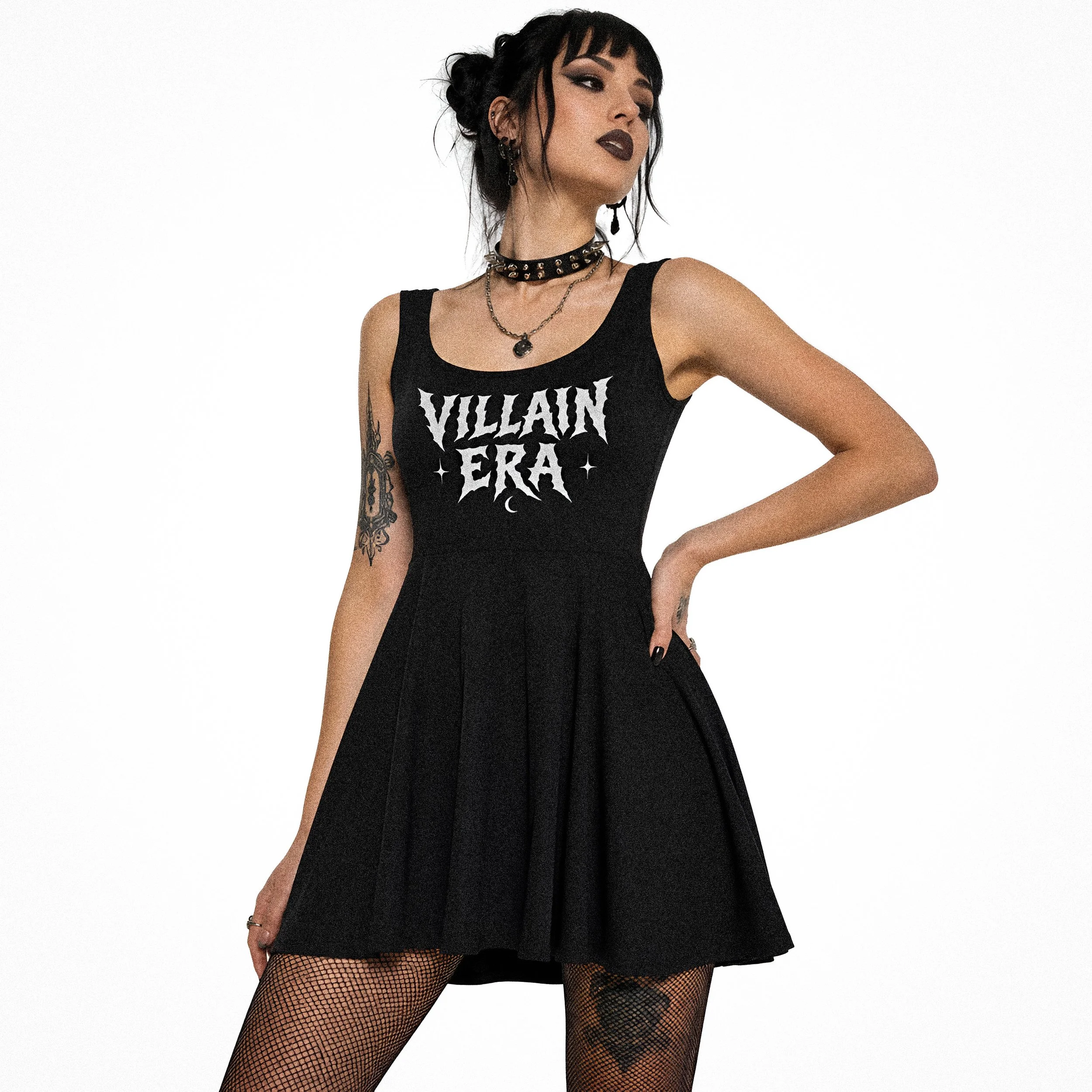 A woman with dark hair styled in a messy bun with bangs, wearing dark lipstick, a black tank top with 'VILLAIN' written on it, fishnet stockings, and layered necklaces, standing against a plain white background.