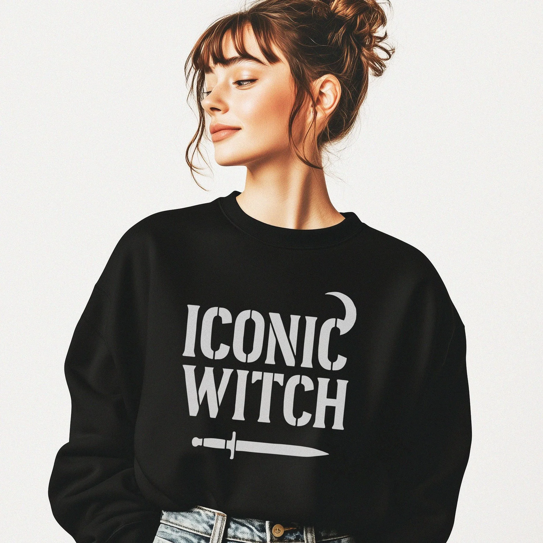 A young woman with brown hair styled in a messy bun and bangs, wearing a black sweatshirt that says 'ICONIC WITCH' with a dagger graphic, standing against a plain background. She is looking to her left with a slight smile.