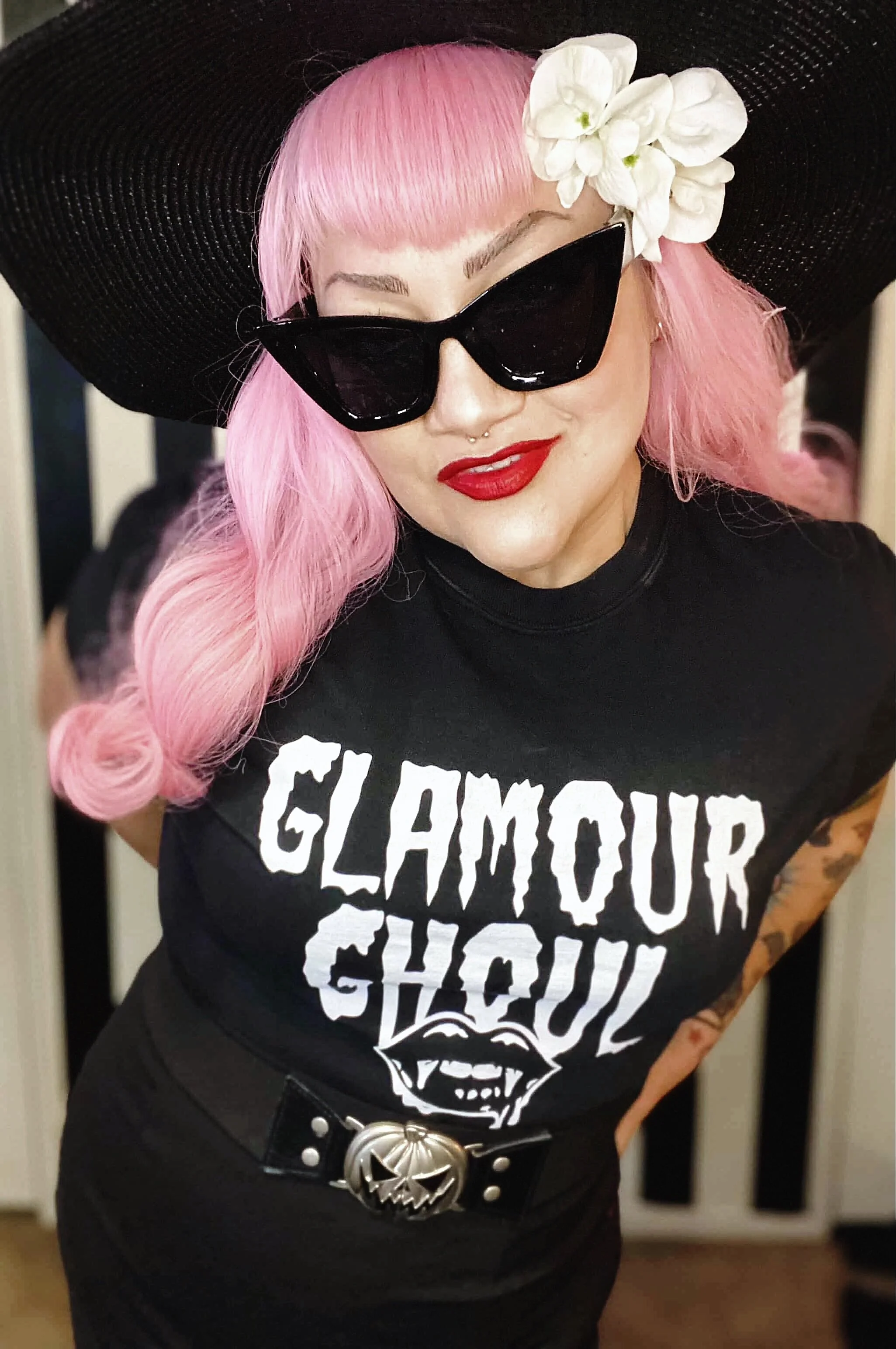 A woman with pink hair wearing black sunglasses, a black wide-brimmed hat with white flowers, a black T-shirt with white graphic text, and a belt with a large decorative buckle, standing indoors.