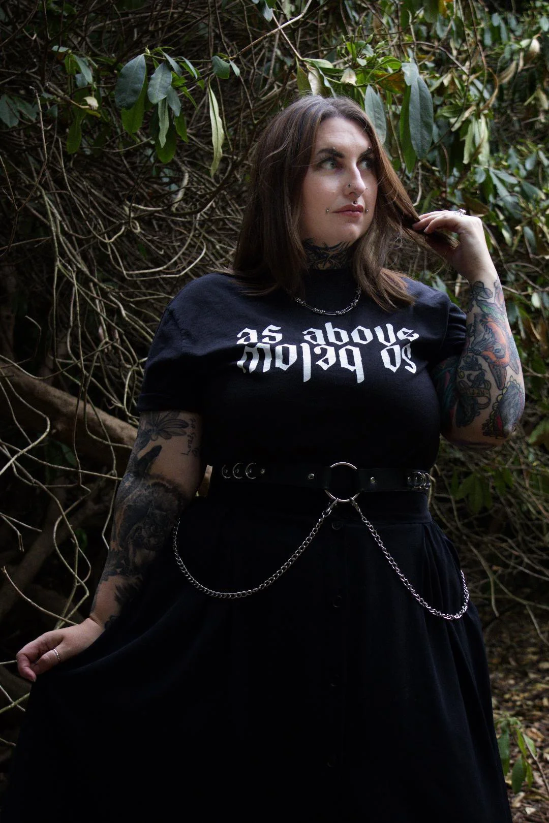 A woman with long brown hair and tattoos on her arms, wearing a black t-shirt with white Gothic-style text, standing outdoors in front of green leafy bushes.