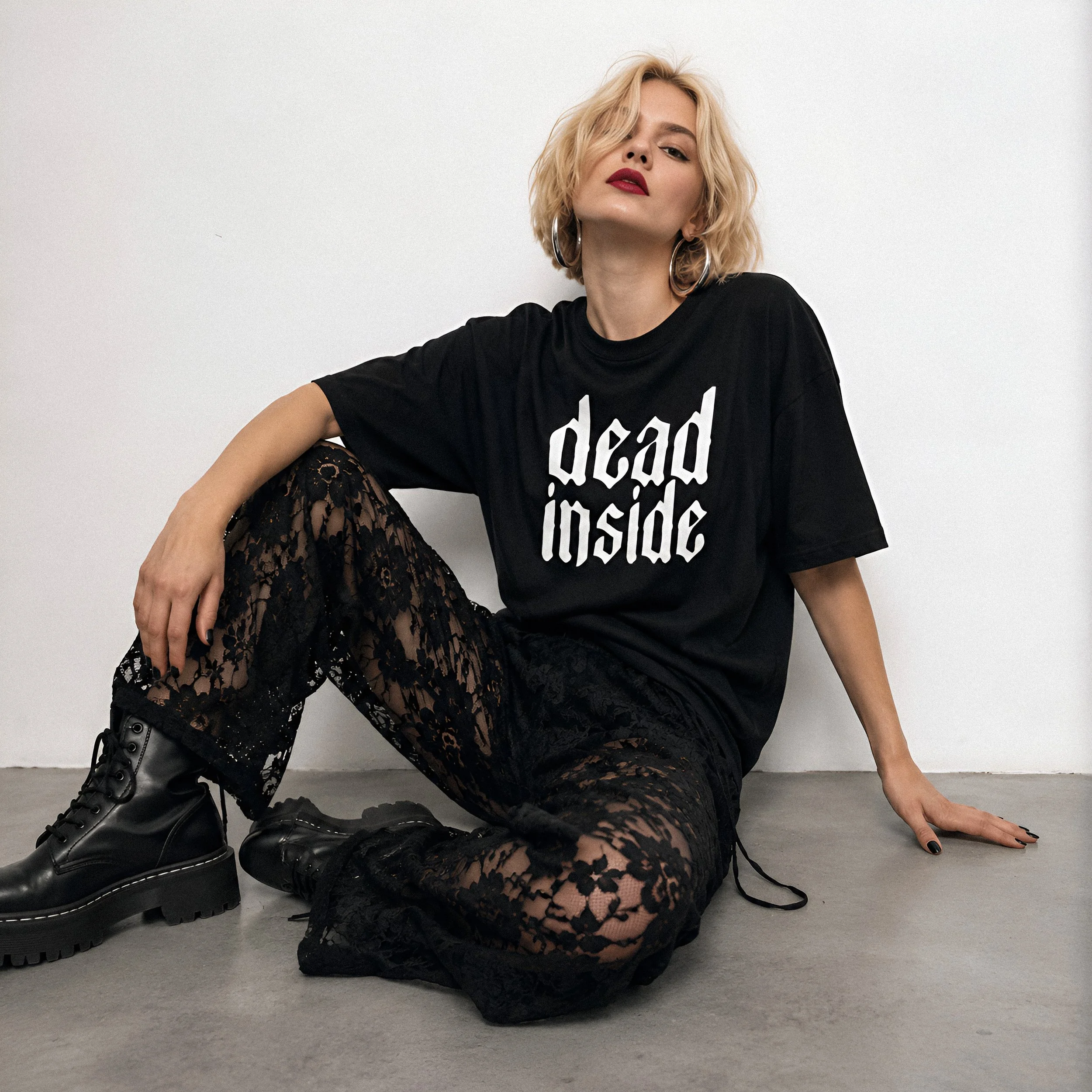 A woman with blonde, wavy hair and red lipstick posing against a plain white wall, wearing a black t-shirt with white lettering and black lace pants, sitting on the ground with one leg crossed and one arm resting on her knee, wearing black combat boots.