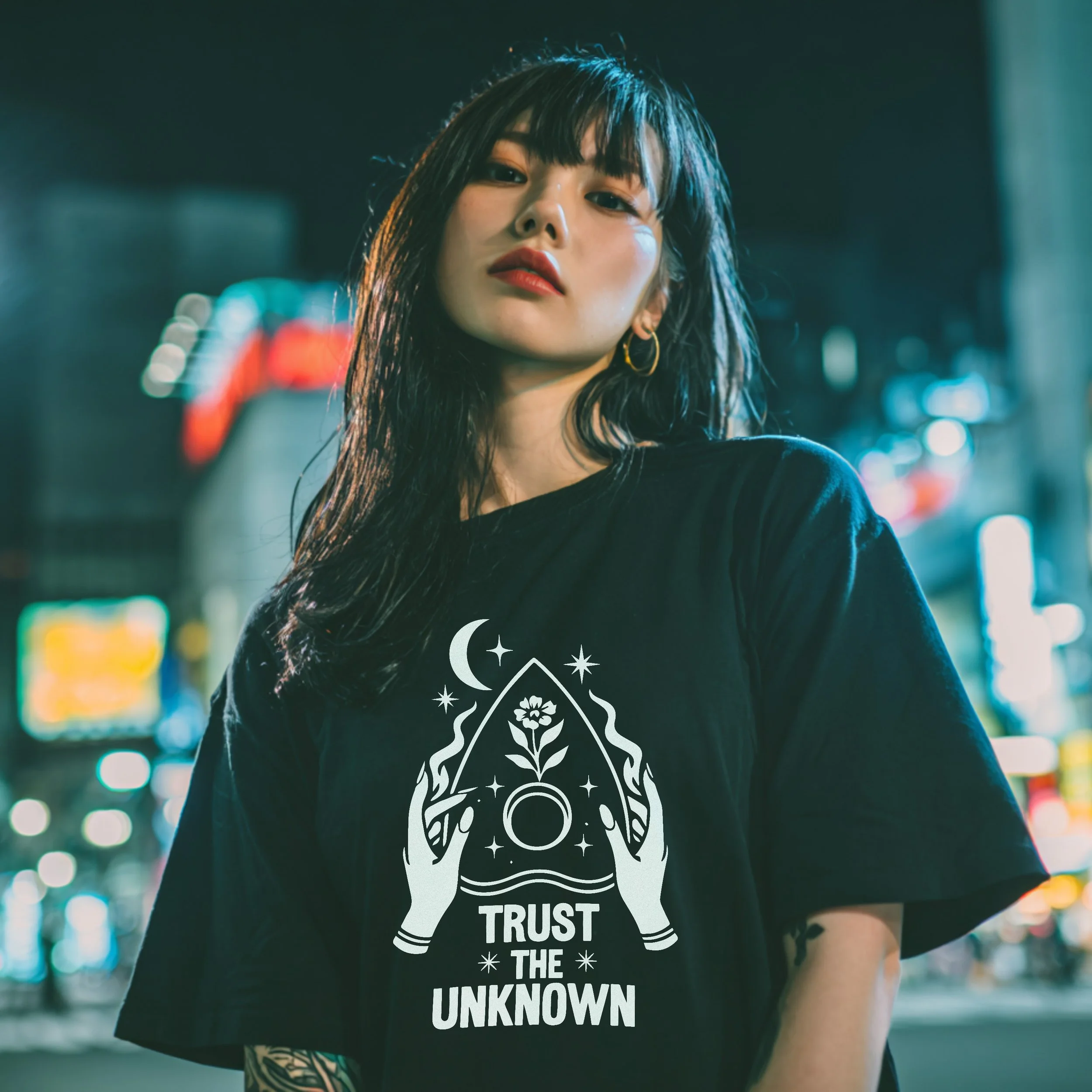 A young woman with black hair and gold hoop earrings wearing a black T-shirt with a graphic design and the text "Trust The Unknown" stands in a city street at night, illuminated by neon lights in the background.