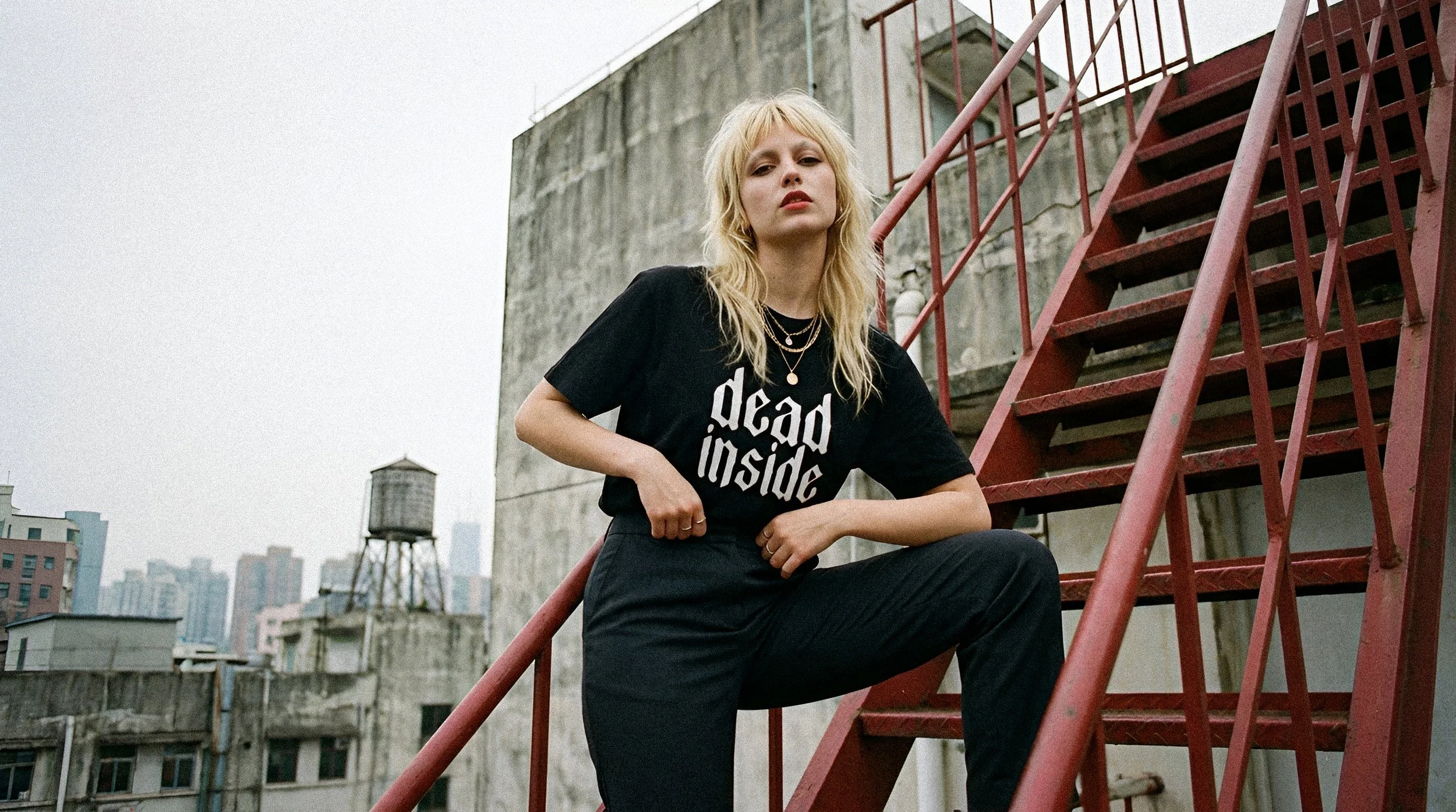 A woman with blonde hair and layered necklaces sitting on a red fire escape ladder on a rooftop in an urban setting.