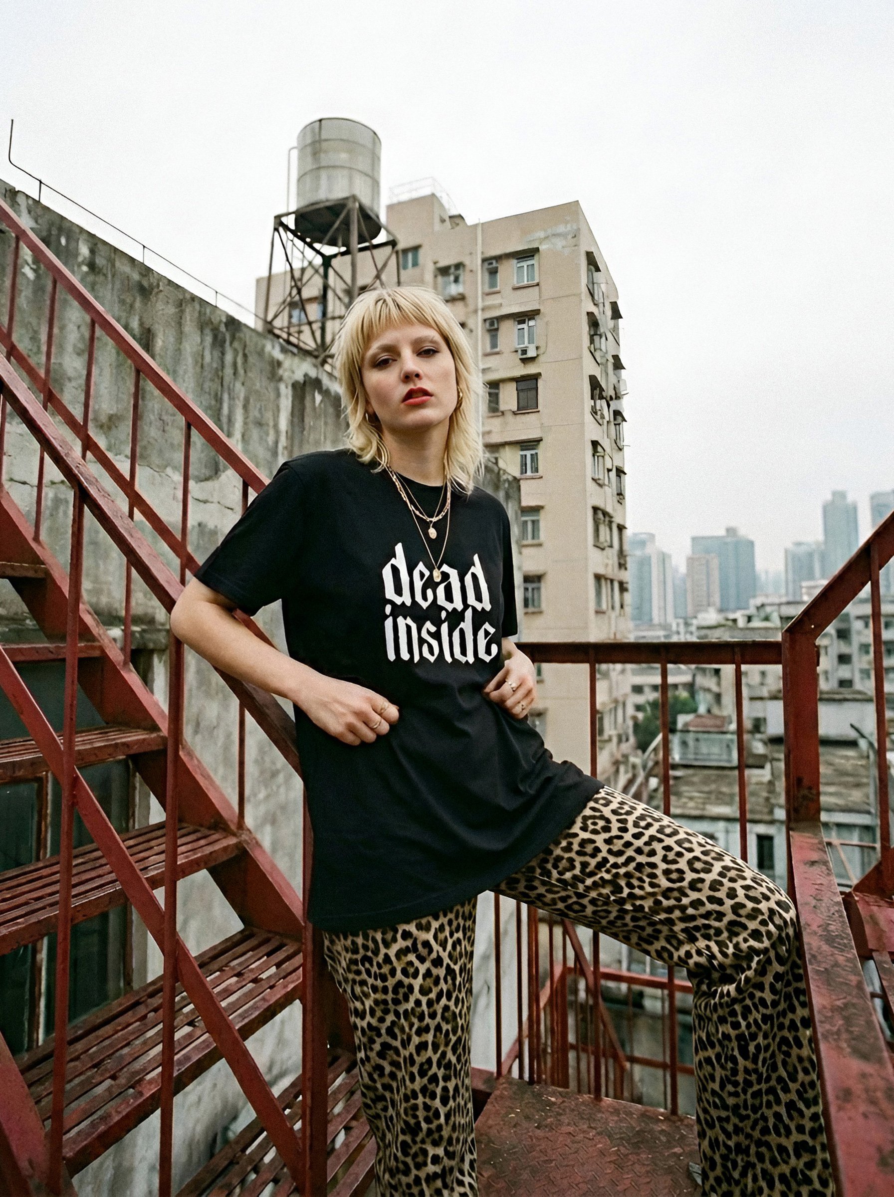 A young woman with blonde hair wearing a black T-shirt with the text 'dead inside' and leopard print pants, standing on a fire escape with city buildings in the background.