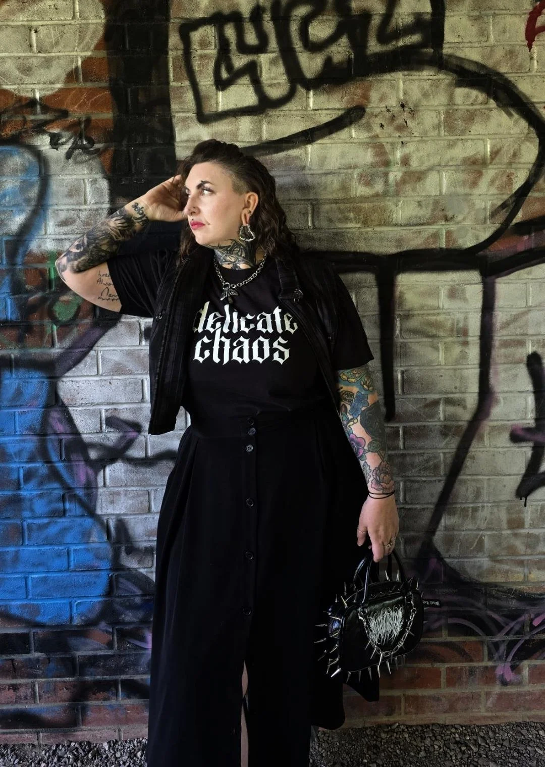 A woman with tattoos on her arms and neck, wearing a black t-shirt with white gothic text, a black skirt, and black accessories, standing in front of a graffiti-covered brick wall.