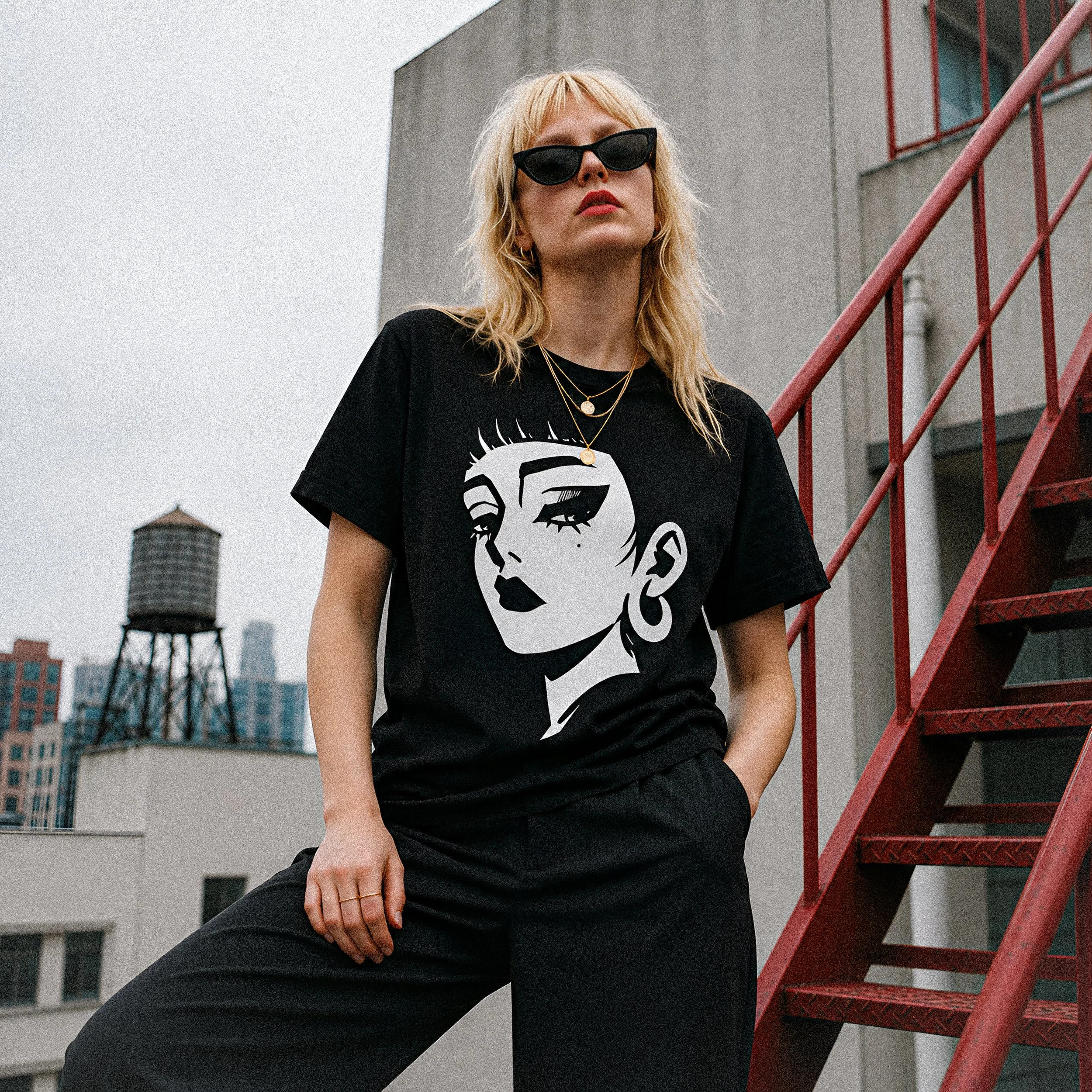 A woman with blonde hair wearing black sunglasses, a black t-shirt with a illustrated face graphic, layered necklaces, and black pants, standing beside an outdoor red metal staircase on a rooftop with tall buildings and water tower in the background.