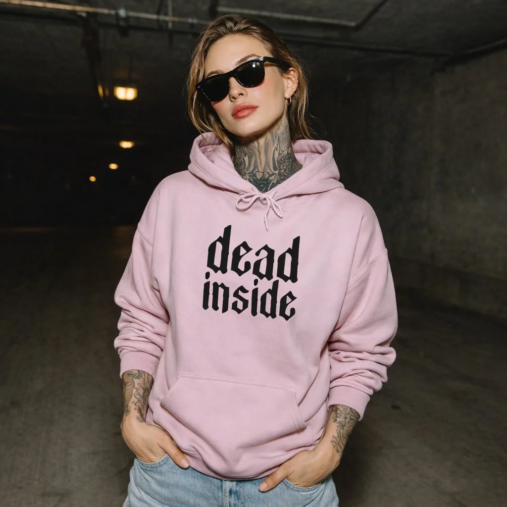 A woman with tattoos on her arms and neck, wearing sunglasses and a pink hoodie with the words 'dead inside' printed on it, standing in a dimly lit underground parking garage.