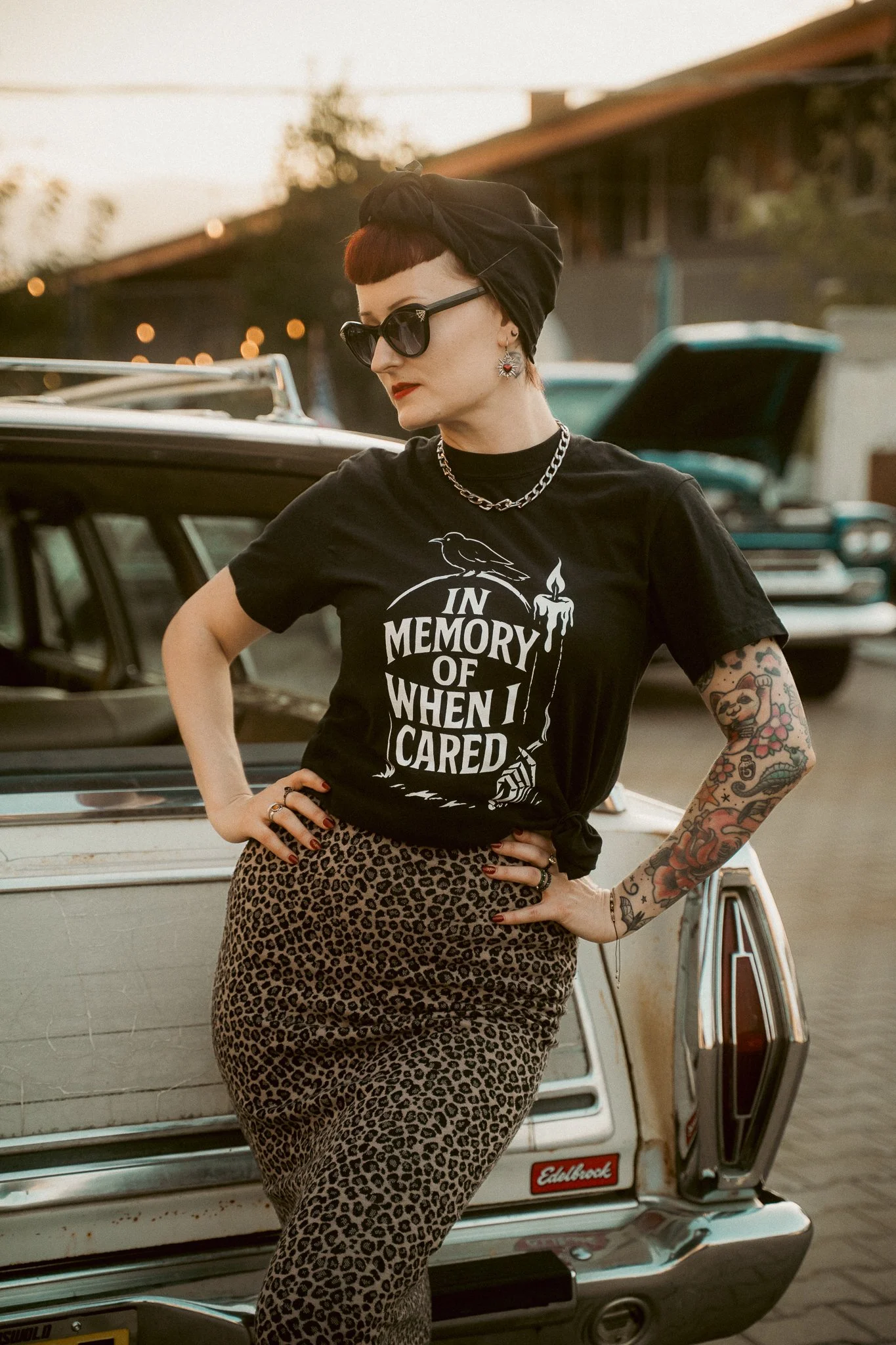 A woman with short red hair wearing black sunglasses, earrings, and a black headscarf, standing in front of a vintage car with an open trunk. She is dressed in a graphic t-shirt and leopard print skirt, with tattoos visible on her left arm.