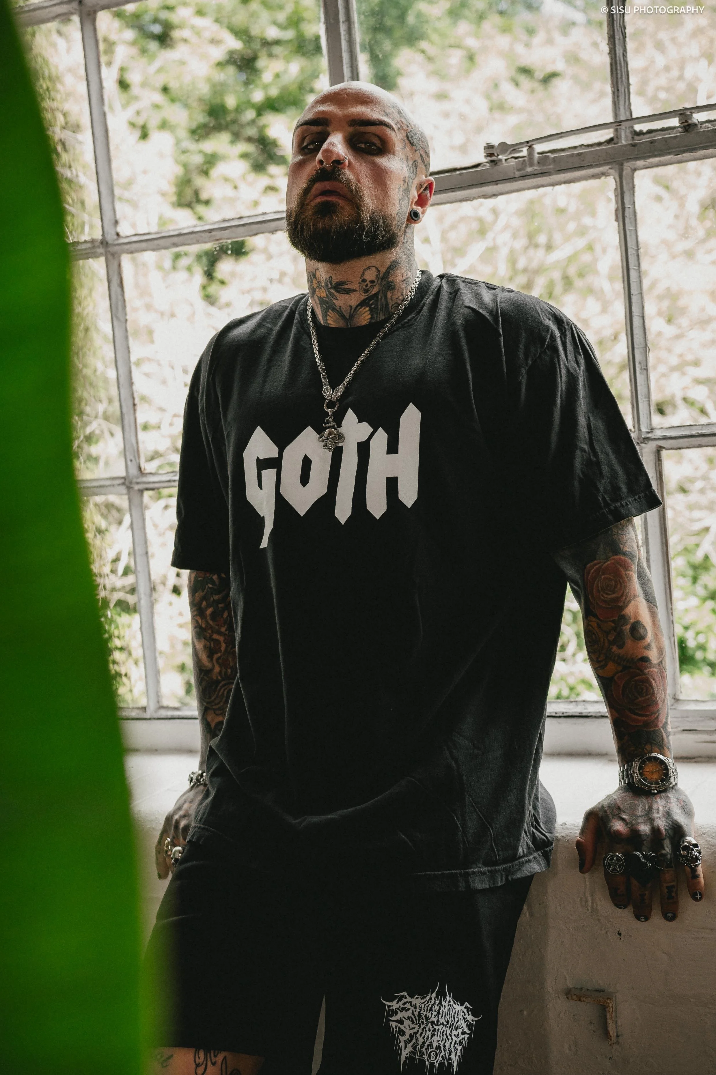A tattooed man with a shaved head, beard, and multiple facial piercings, wearing a black T-shirt with the word 'GOTH' printed on it. He is standing inside near a large window with a metal grid, outdoors with greenery visible in the background.
