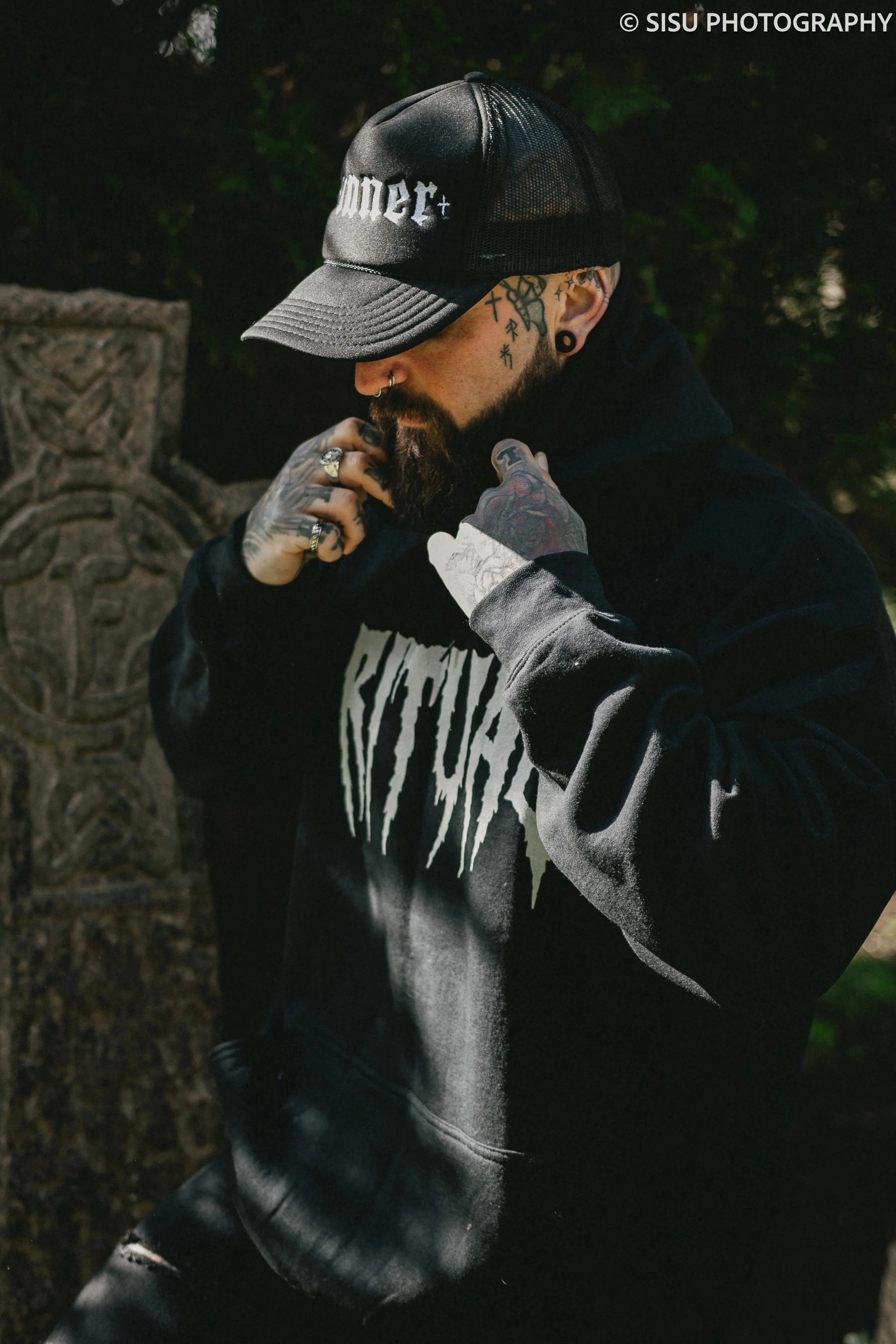 A tattooed man wearing a black baseball cap and black hoodie, with a beard and piercings, standing outdoors near a stone monument or sculpture, with trees and greenery in the background.