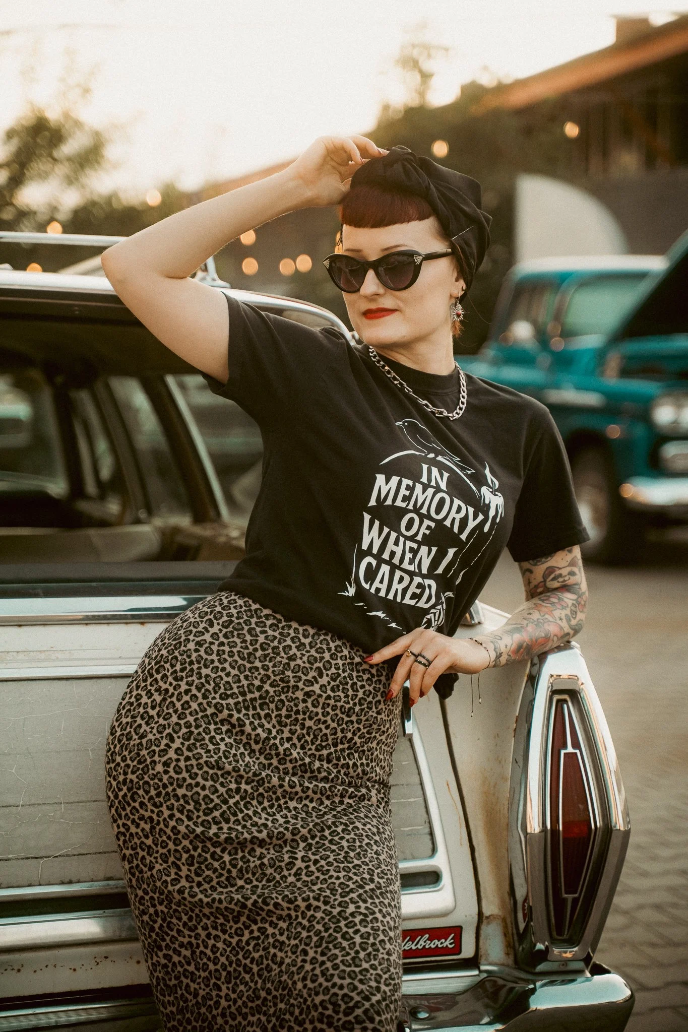 A woman with short red hair, sunglasses, and tattoos wearing a black shirt and leopard print skirt leaning against a white vintage car with an open door.