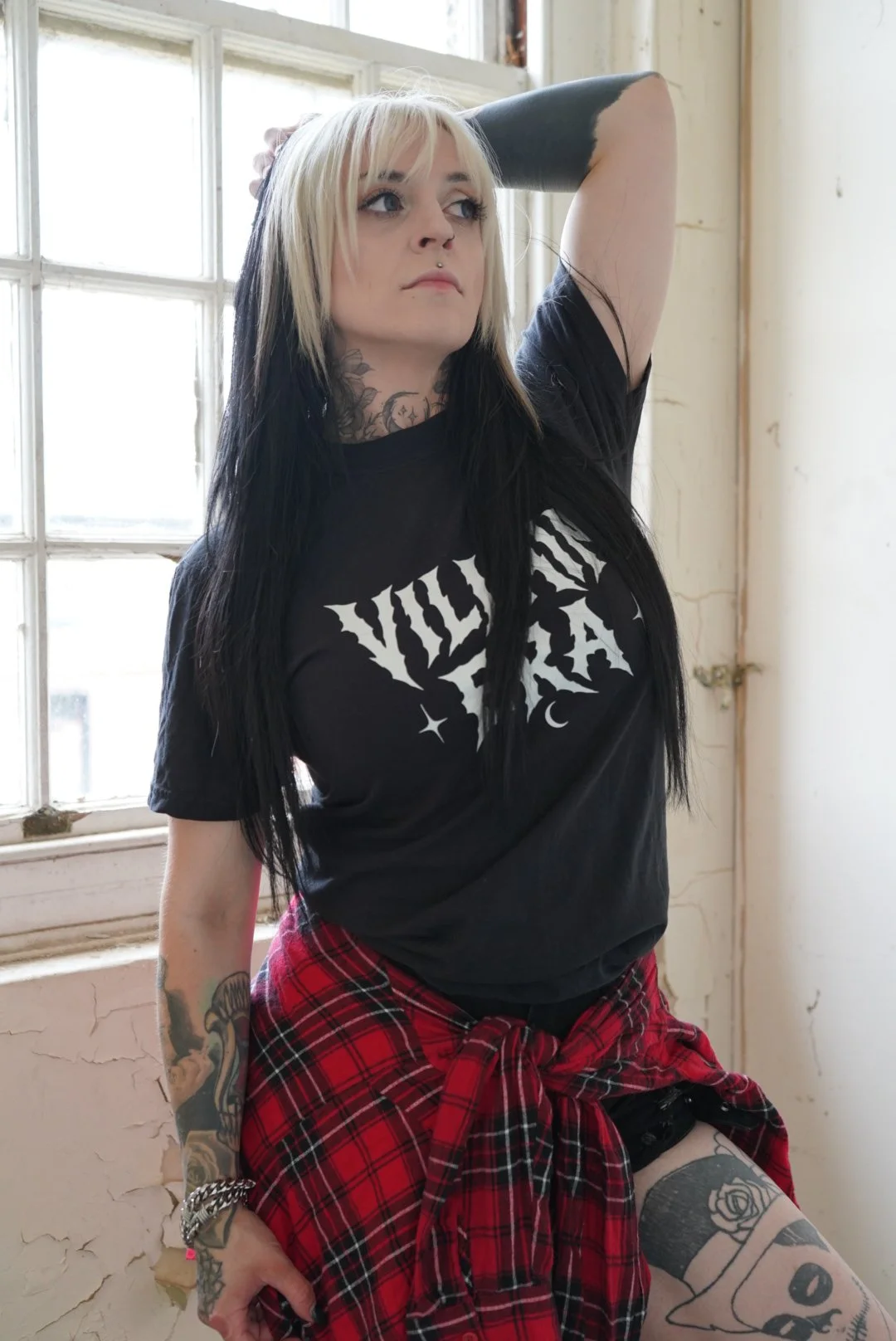 A woman with blonde and black hair, tattoos on her left arm and thigh, wearing a black band t-shirt with white text and a red plaid shirt tied around her waist, standing by a window with peeling paint and cracked walls.