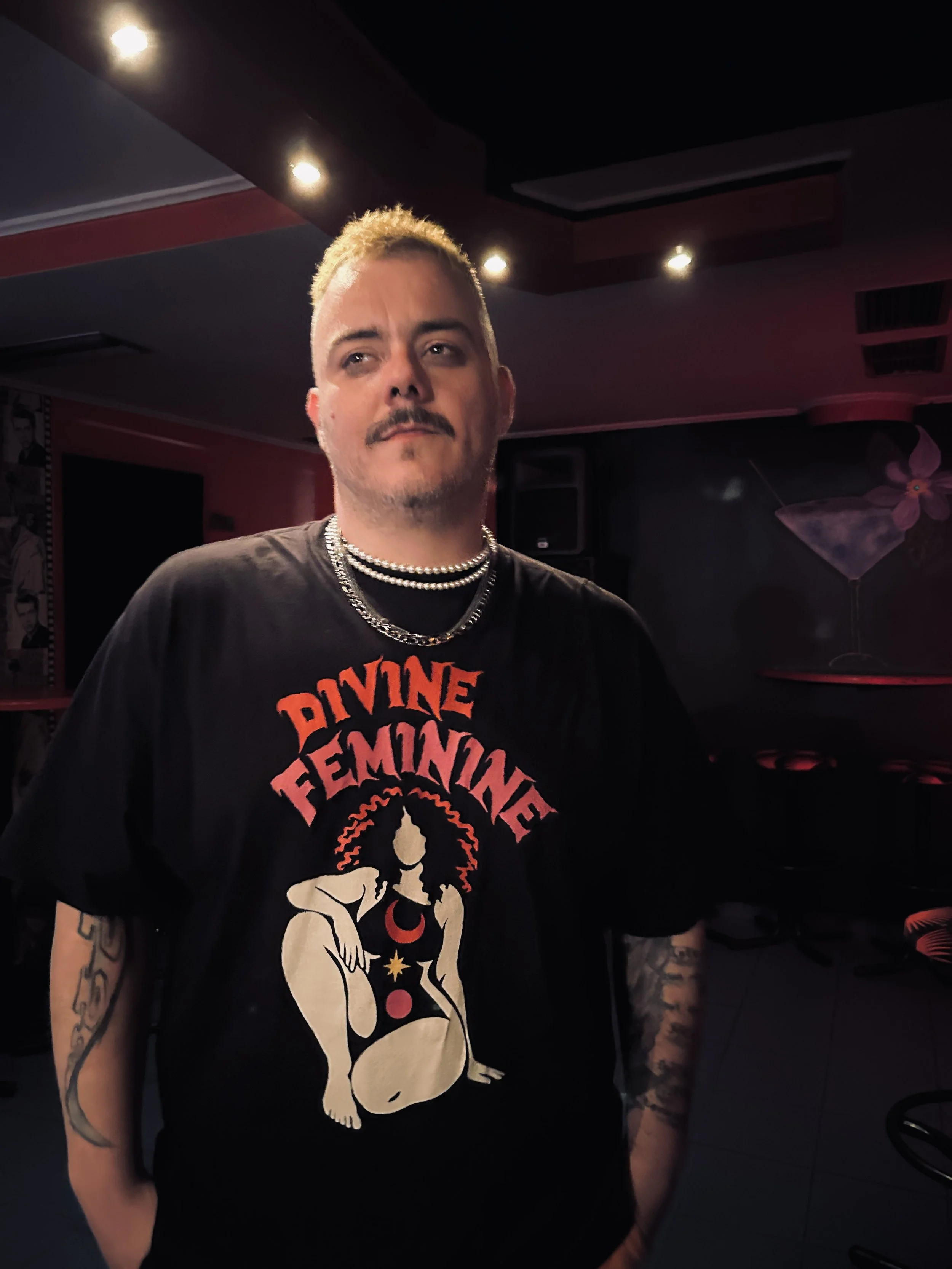 A young man with short, bleached blonde hair and a goatee, wearing a black T-shirt with a red and beige graphic, a pearl necklace, and multiple tattoos on his arms, stands in a dimly lit room with a pink and black decor, including a poster on the wall and a large pink flower decoration.