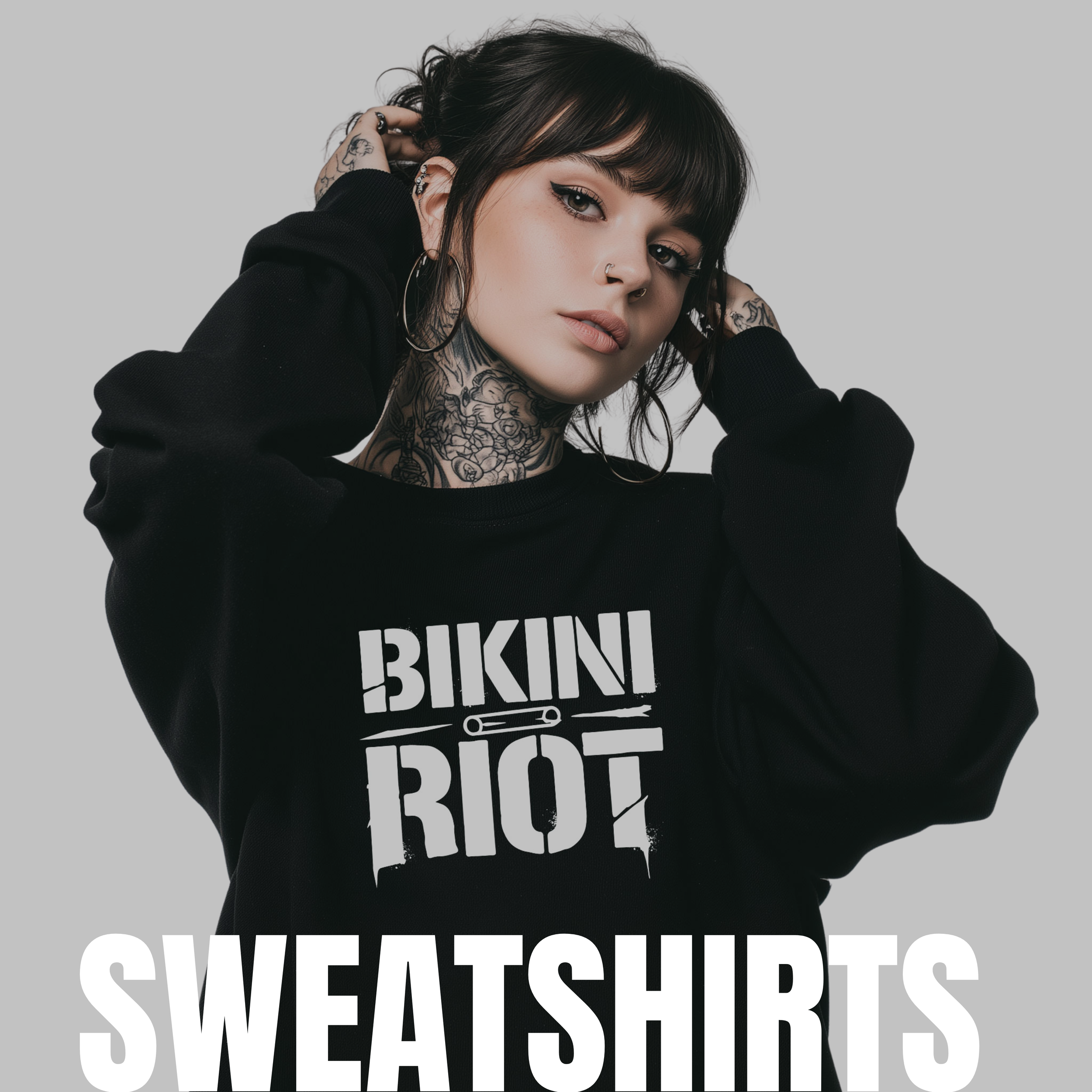 A young woman with tattoos, dark hair, and hoop earrings wearing a black sweatshirt with the text 'Bikini Riot' and holding her hair, against a plain gray background.