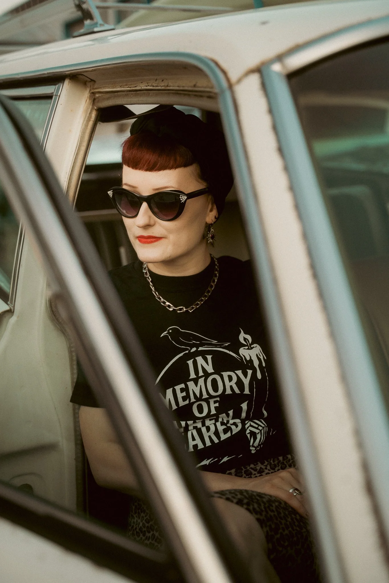 A woman with short red hair wearing sunglasses, earrings, a chain necklace, a black t-shirt with white text and graphic, and leopard print pants, sitting inside an old car.