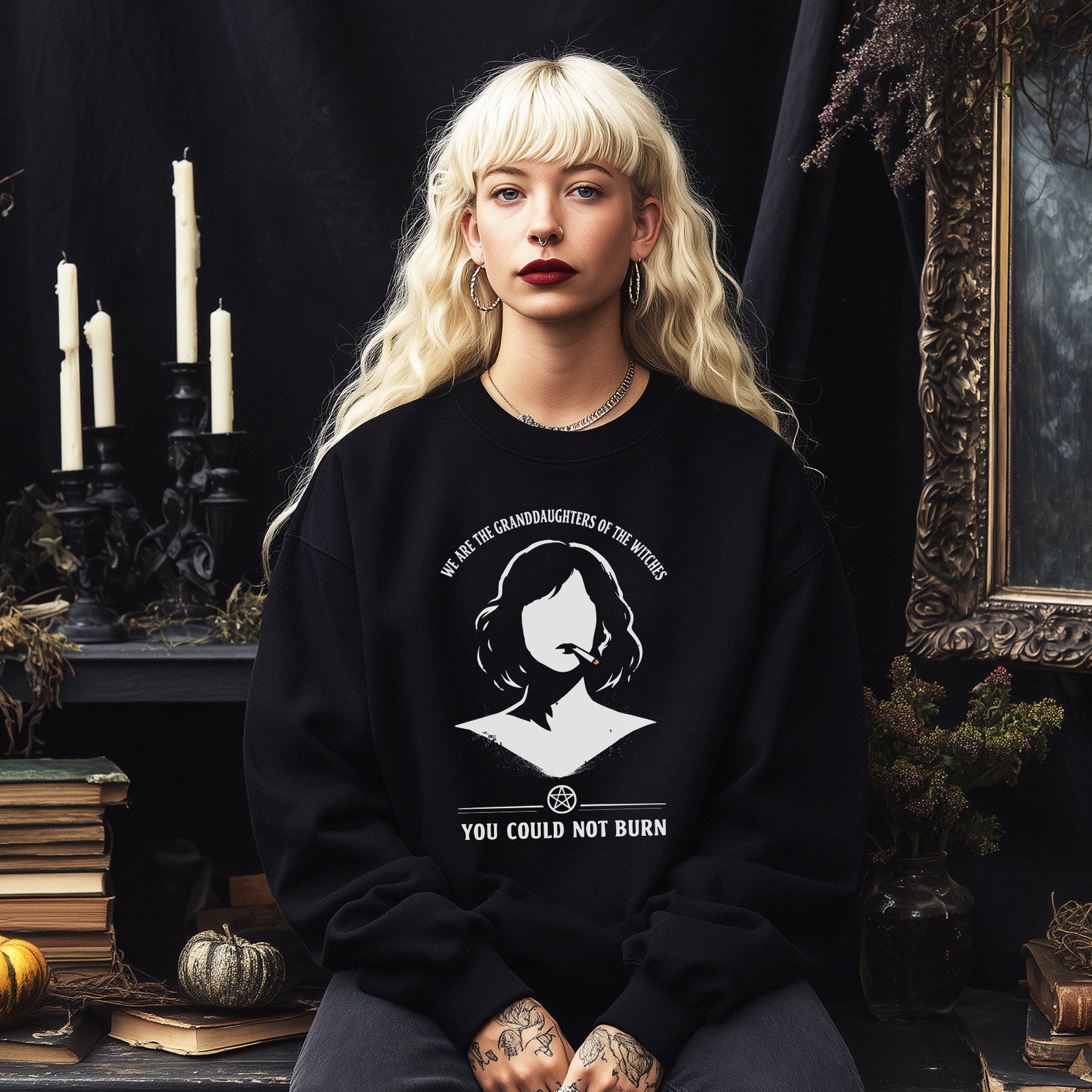 A young woman with long blonde wavy hair and tattoos on her hands, wearing a black sweatshirt with a graphic of a woman's silhouette, text that reads 'We are the granddaughters of the witches you could not burn,' and accessories like hoop earrings and a choker, sitting in a dark gothic-themed room with candles, dried plants, books, and a large ornate mirror in the background.