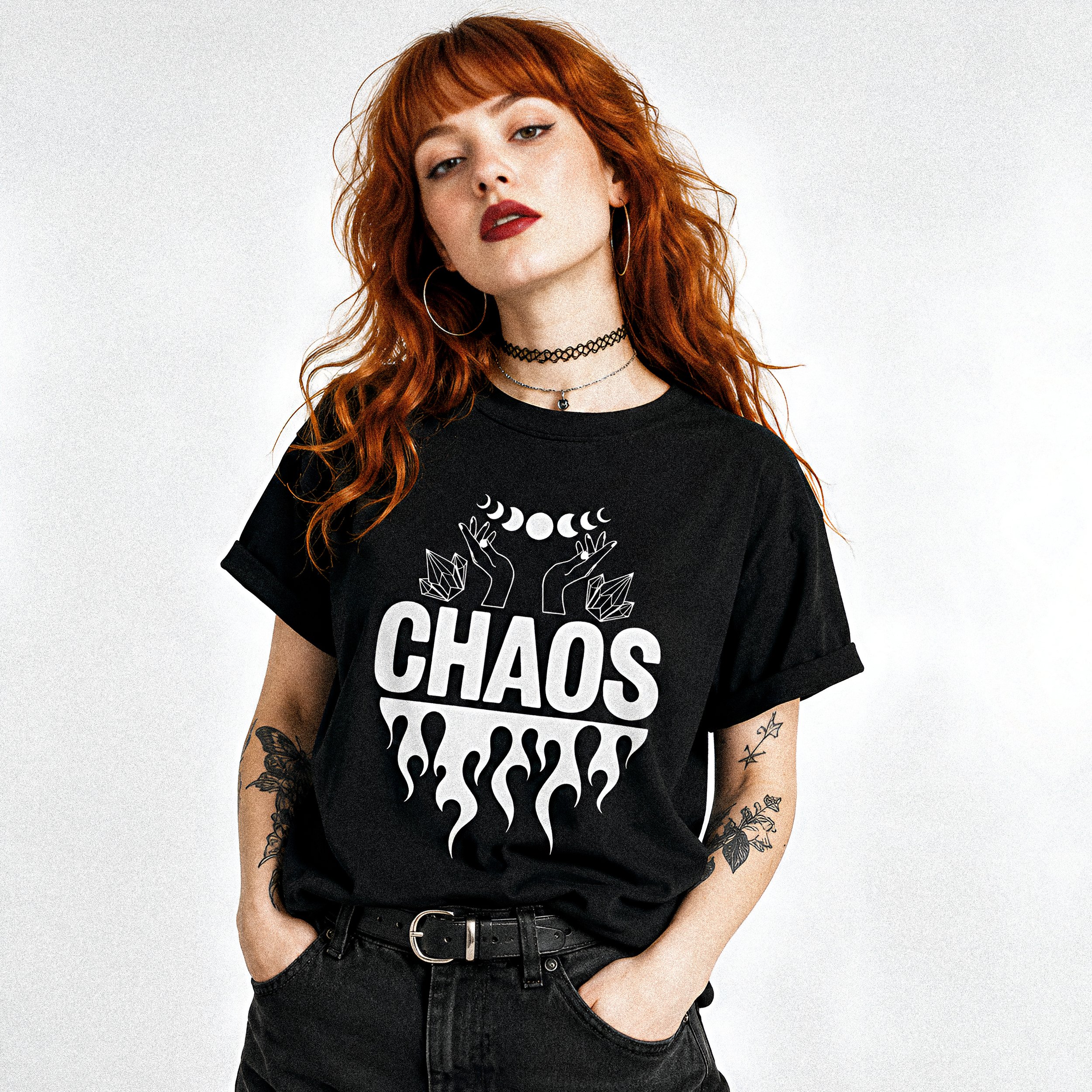 A woman with red hair, tattoos on her arms, wearing a black t-shirt with white graphics, multiple necklaces, and hoop earrings, standing against a plain white background.