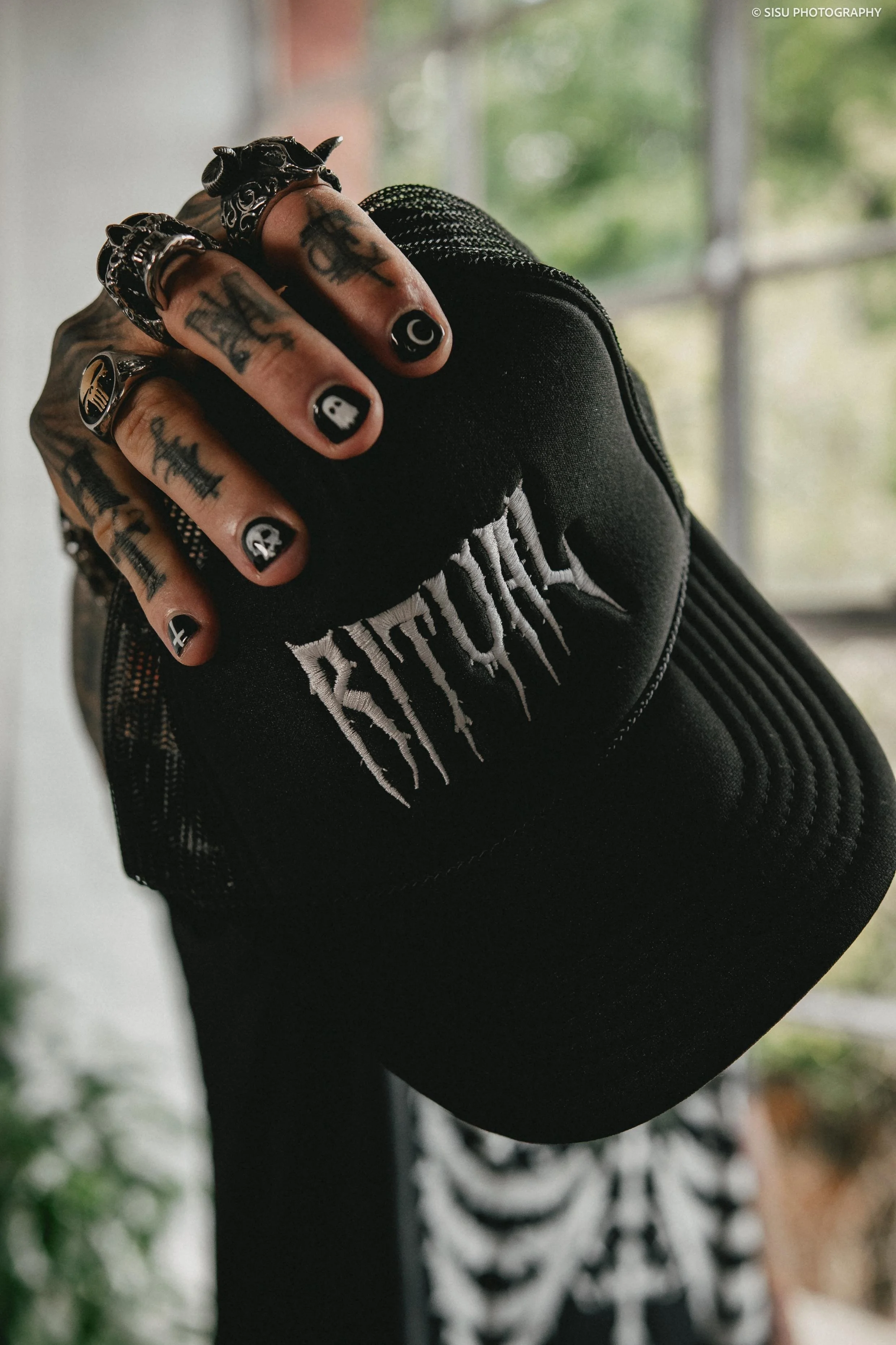 Close-up of a hand with tattoos and multiple rings, holding a black baseball cap with white embroidered text, with a window and blurred background.