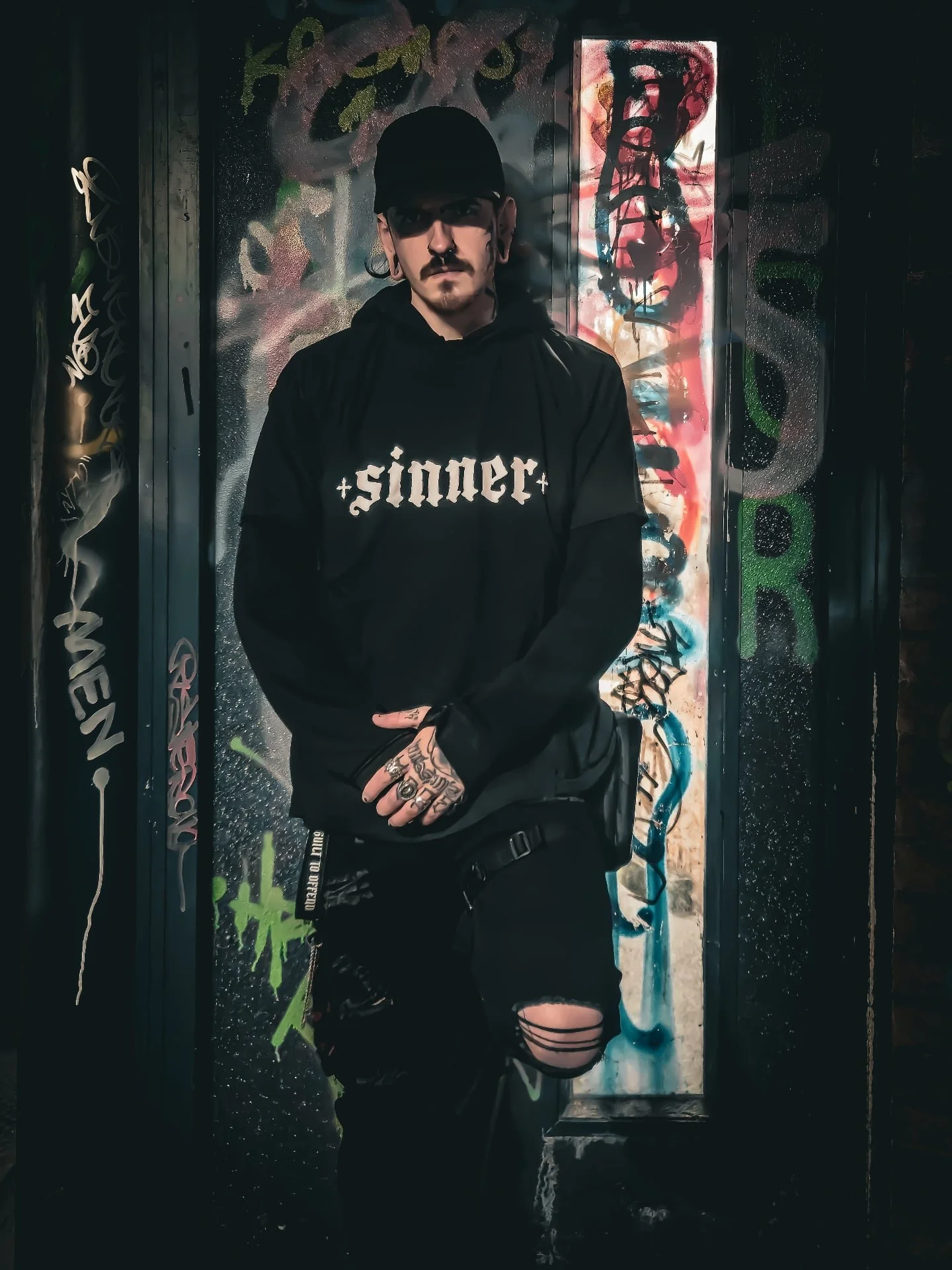 Young man with tattoos, wearing a black hoodie with 'sinner' written on it, standing against a colorful graffiti wall in a dark setting.