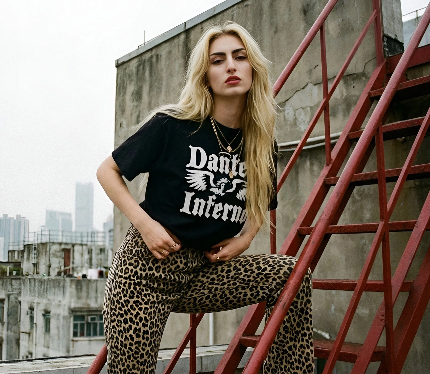 A young woman with long blonde hair poses on a rooftop with city buildings in the background. She is wearing a black t-shirt with white text and a graphic, leopard print pants, and layered gold necklaces. She is standing next to a red metal staircase attached to a weathered concrete wall.