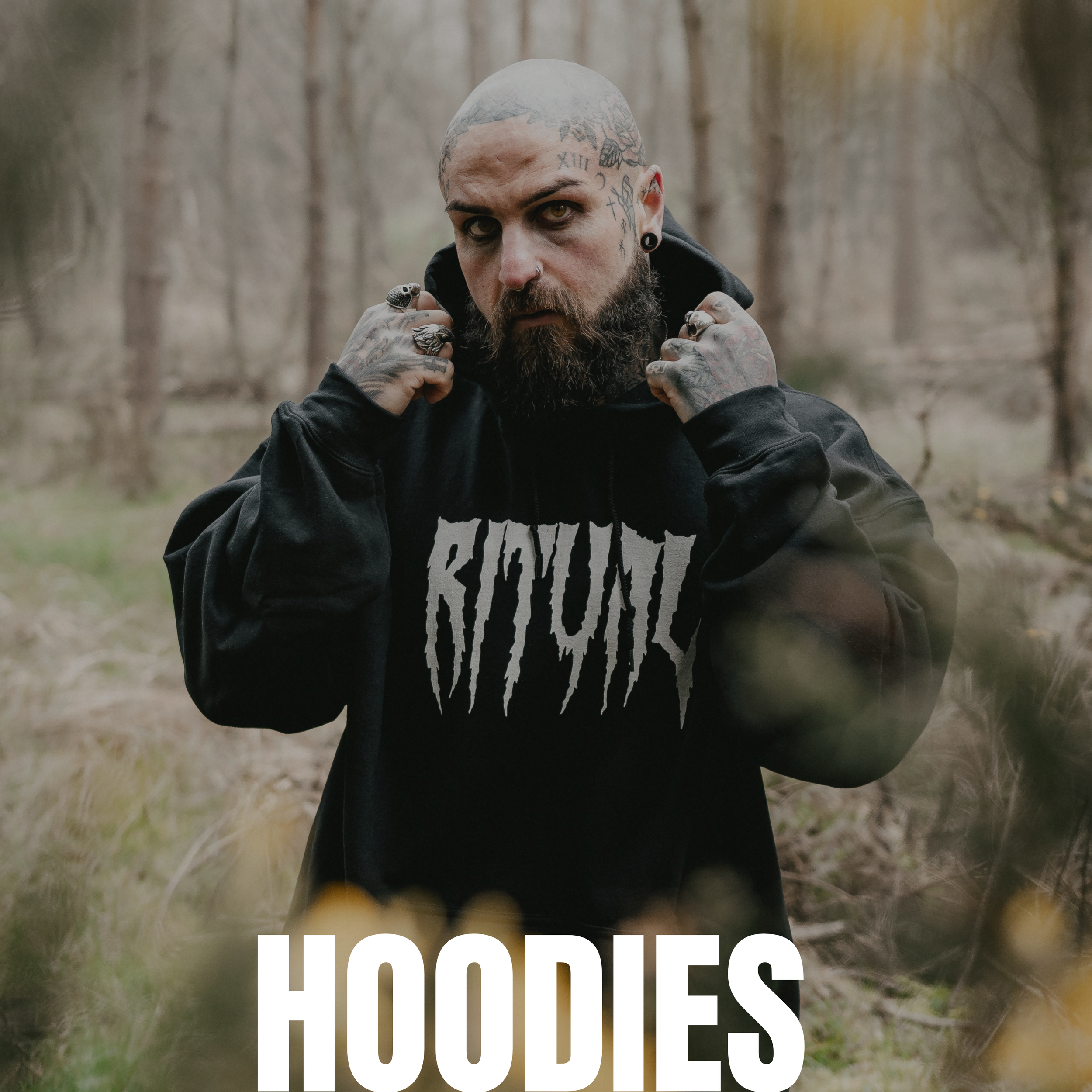 A man with tattoos and facial piercings wearing a black hoodie with the word 'RITUAL' in white, standing in a forest surrounded by trees and foliage.