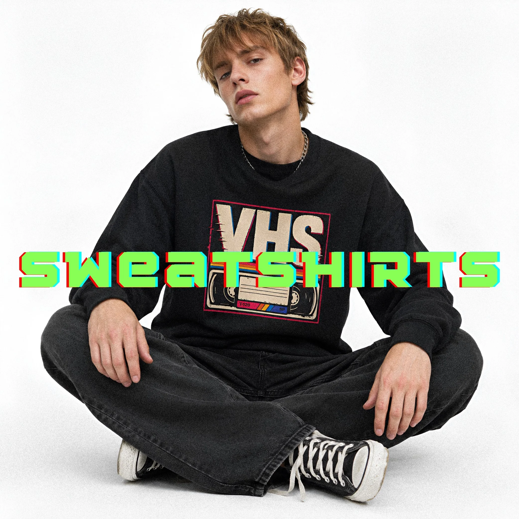 A person sitting on the floor with crossed legs, wearing a black shirt with multicolored text, black jeans, and white sneakers, against a white background.