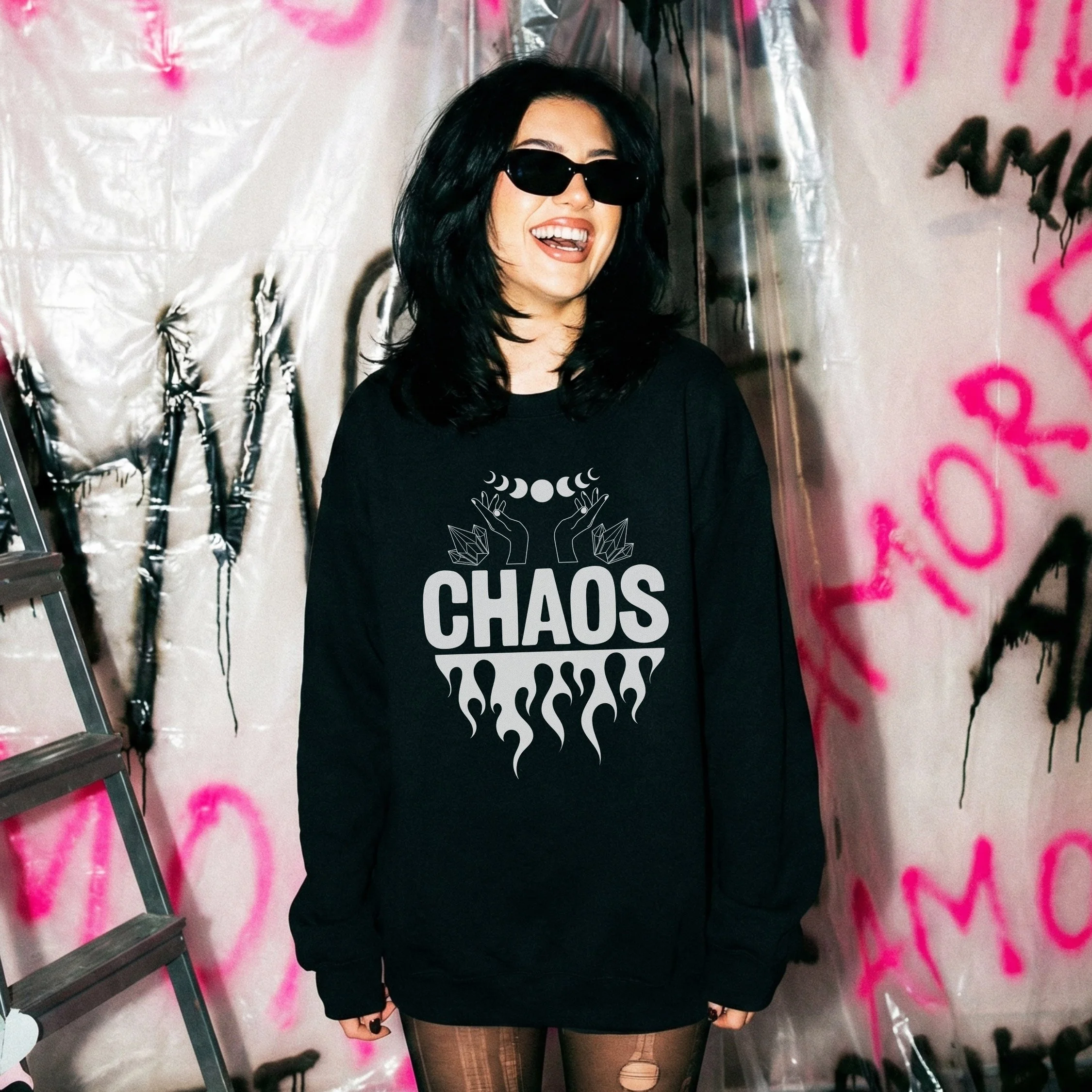 A woman with black hair and sunglasses wearing a black sweatshirt with the word "CHAOS" and a graphic of moon phases and flames. She is smiling and sticking her tongue out, standing in front of a graffiti-covered wall.